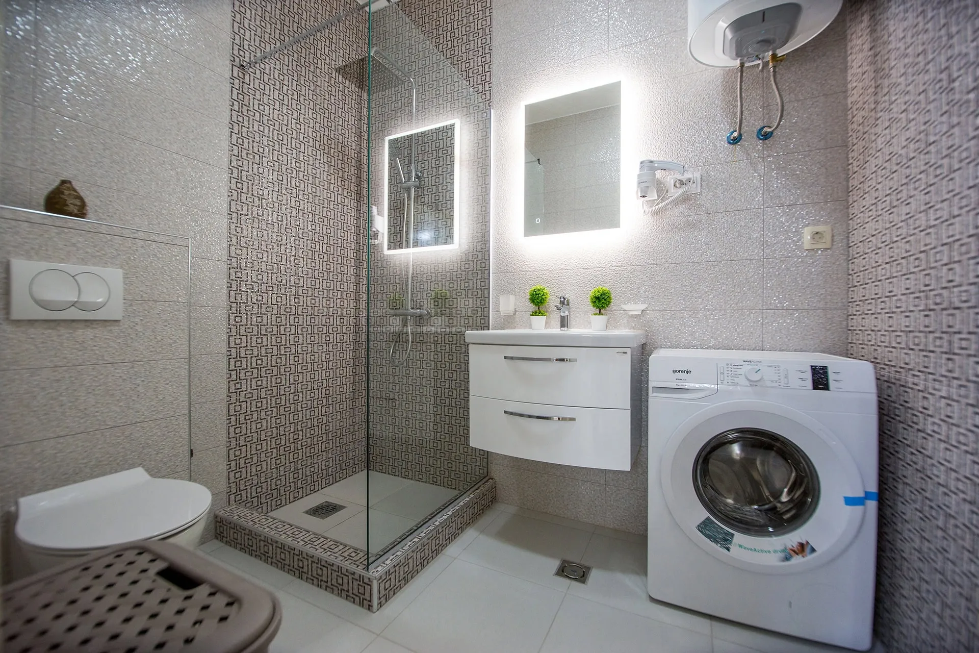 Bathroom featuring a glass shower, toilet, vanity with mirror, and a washing machine on patterned tiles.