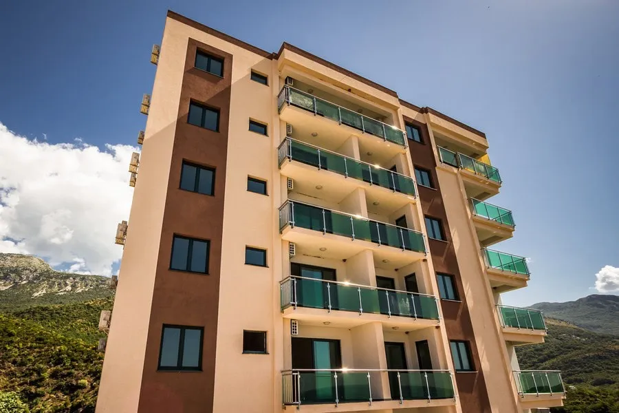 Exterior view of a multi-story residential building with balconies against mountains and blue sky.