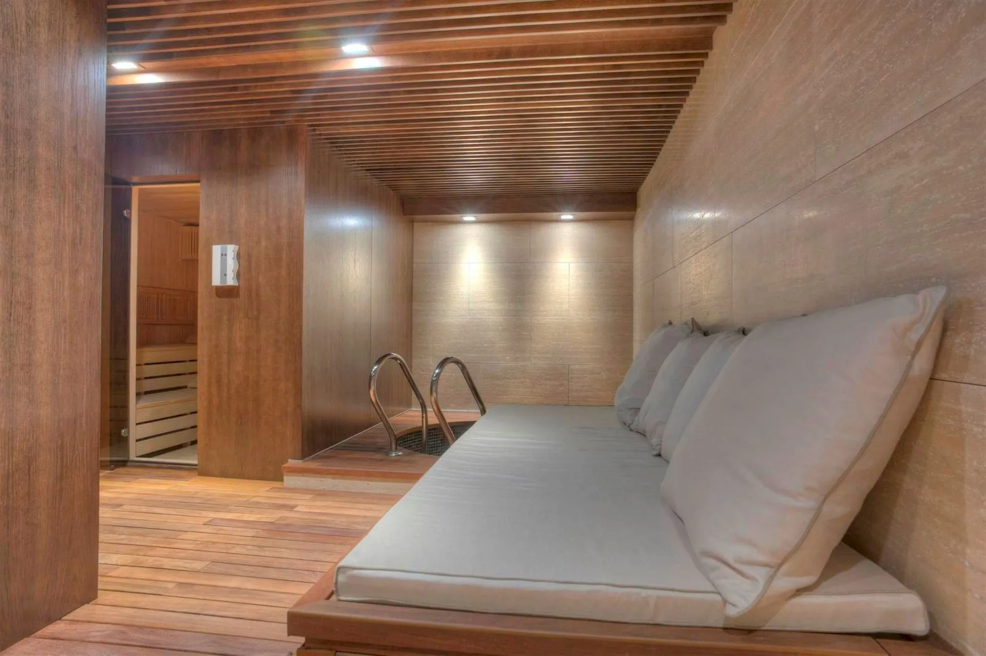 Indoor relaxation area with a cushioned lounge bench next to a pool ladder and sauna entrance.