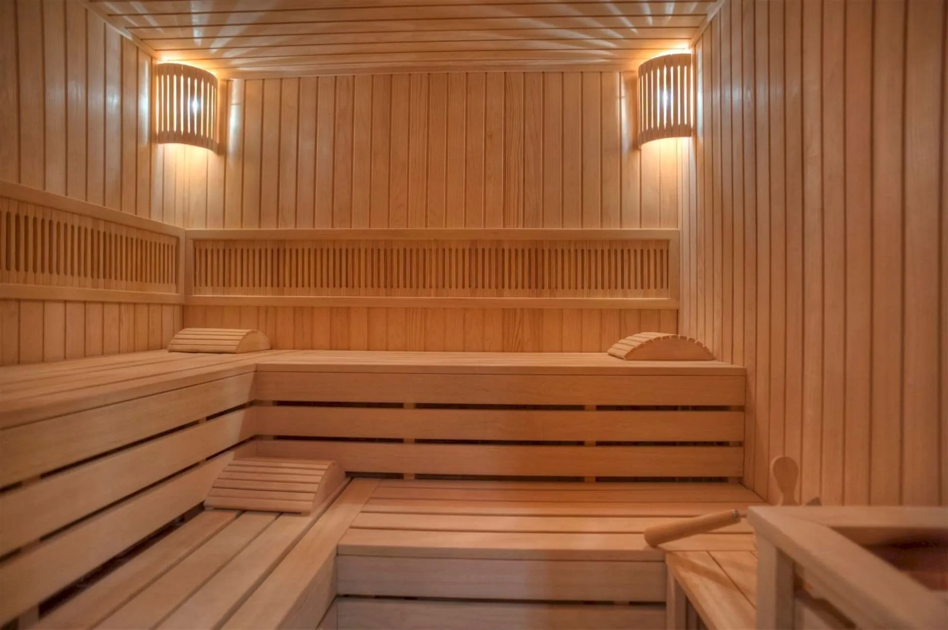Interior of a wooden sauna featuring tiered benches, slatted walls, and warm ambient wall lighting fixtures.