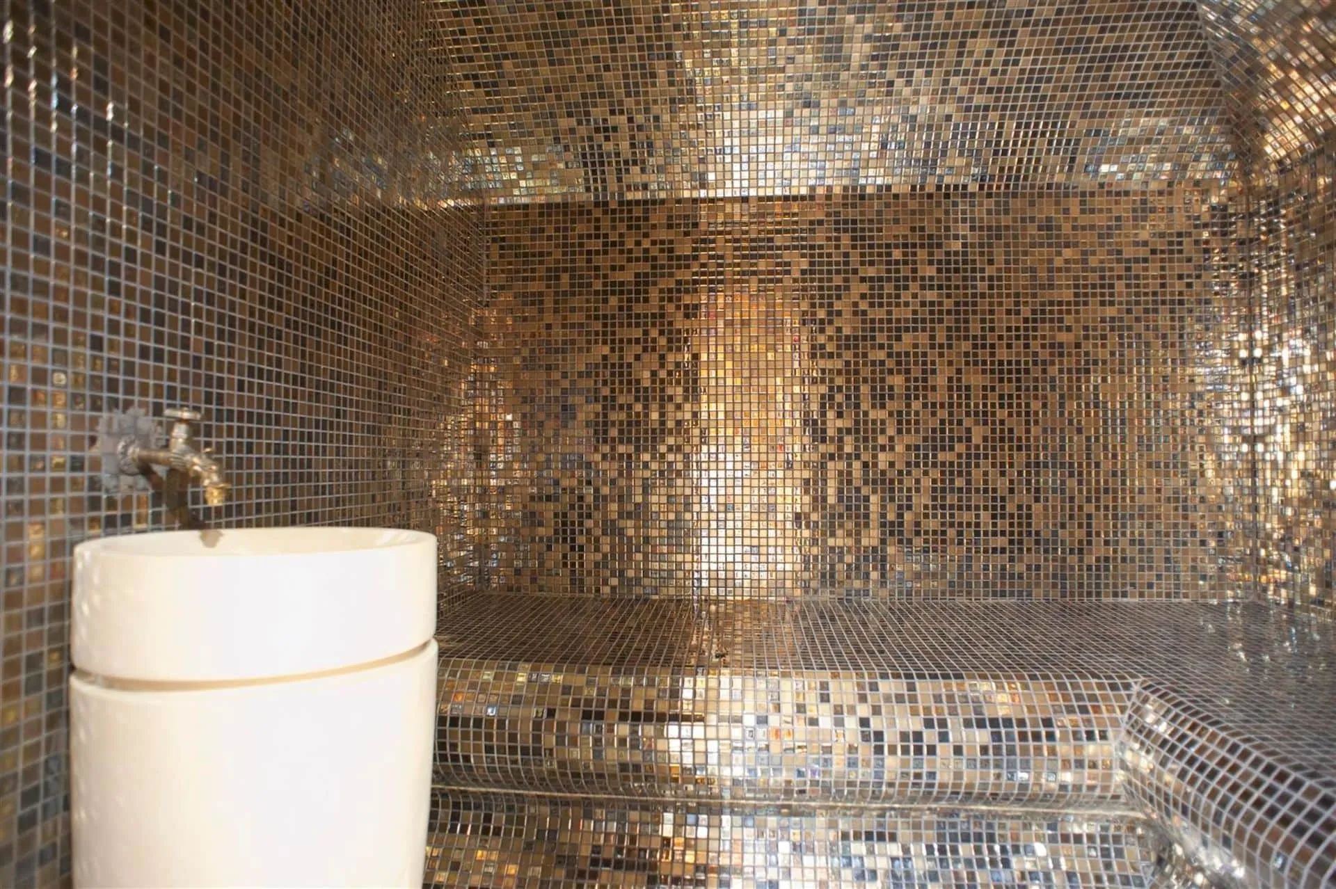 Bathroom with gold mosaic tiles on walls and ceiling, featuring a white cylindrical vessel sink and brass faucet.