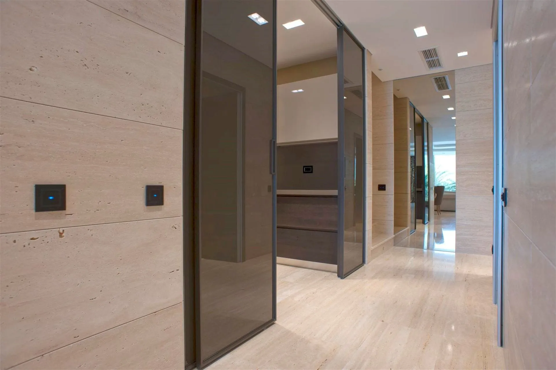 Modern hallway with light marble floors and walls, sliding glass doors, recessed lighting, and wall control switches.