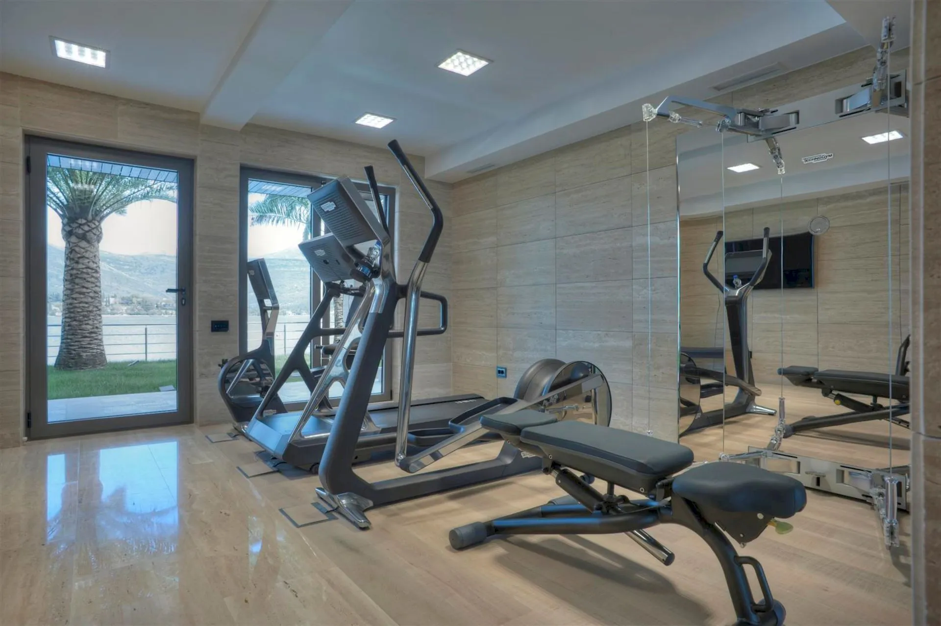 Home gym with a treadmill, elliptical trainer, and weight bench. Features a large mirror wall and glass doors leading to an outdoor view.
