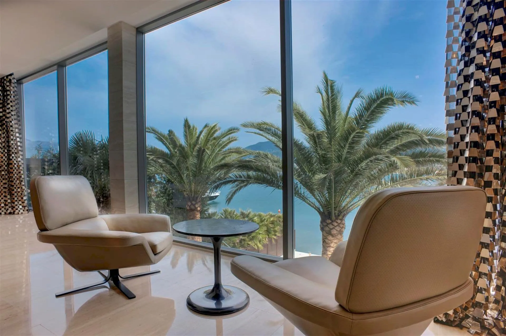 Bright room with two beige armchairs and a small table in front of floor-to-ceiling windows overlooking the sea and palm trees.