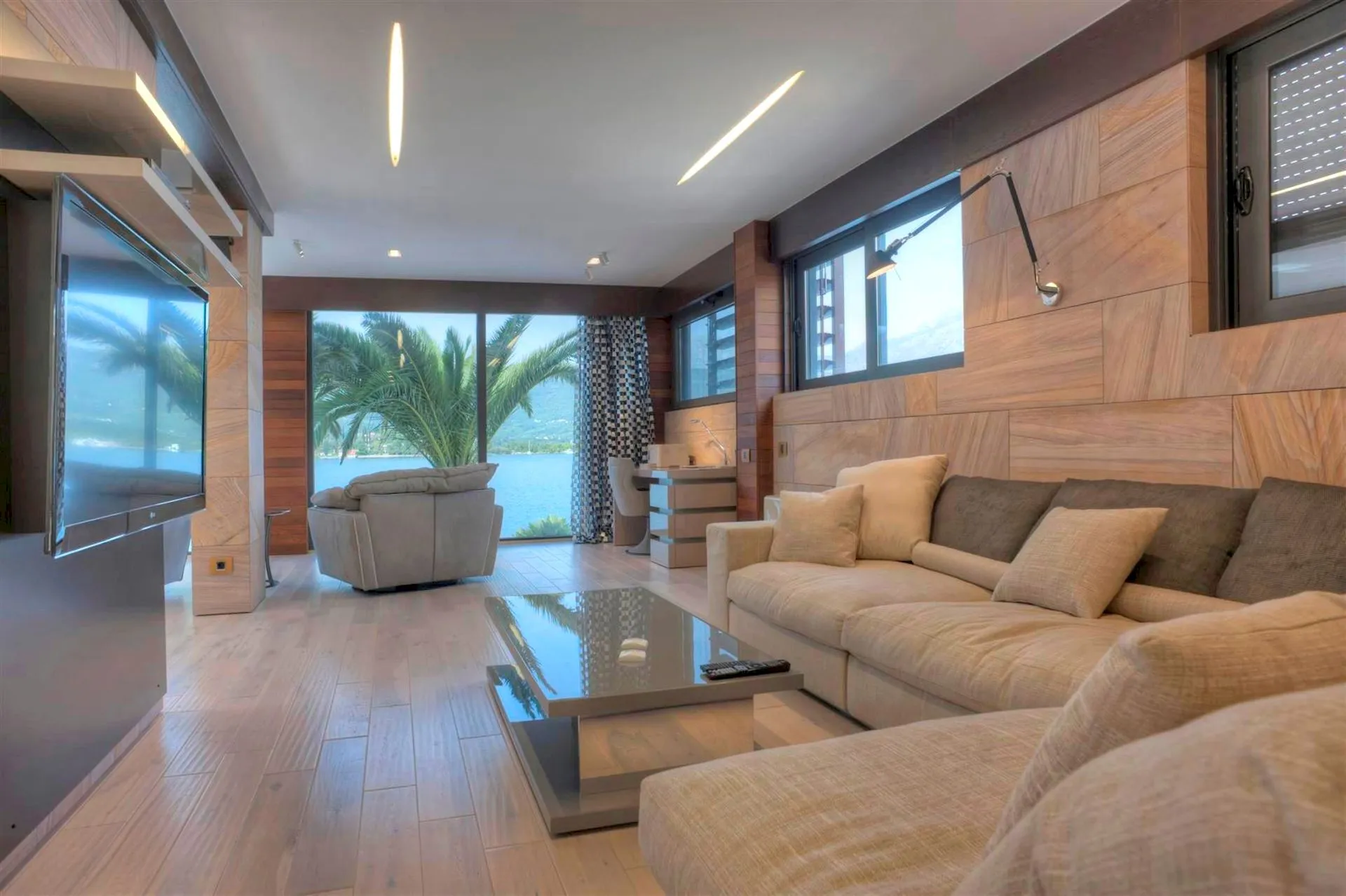 Modern living room with beige sofa, TV unit, glass table, and water view.
