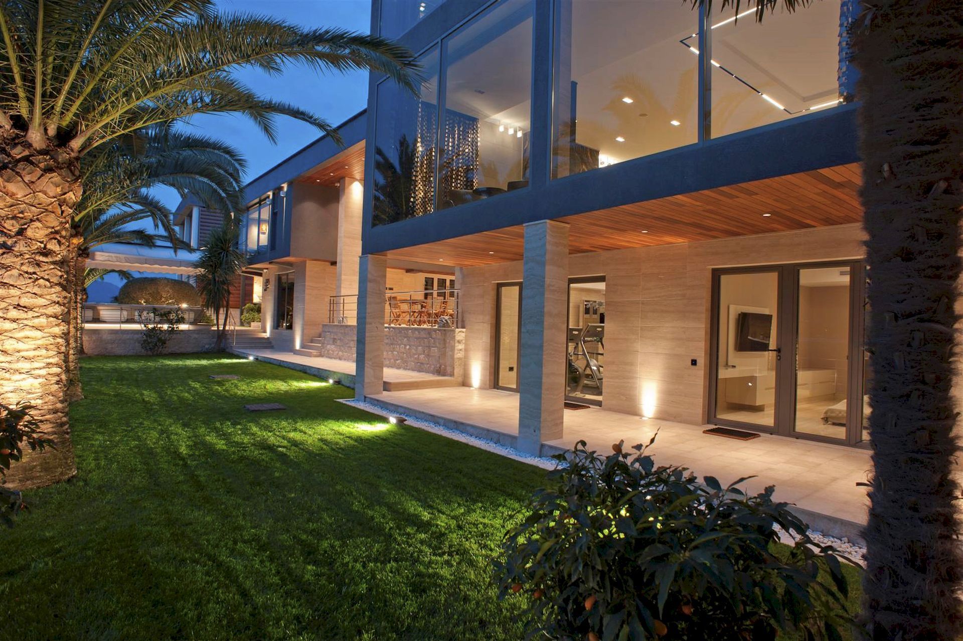 Modern villa in Krašići, Montenegro: panoramic glazing, a lit terrace, and a well-kept garden with palm trees.