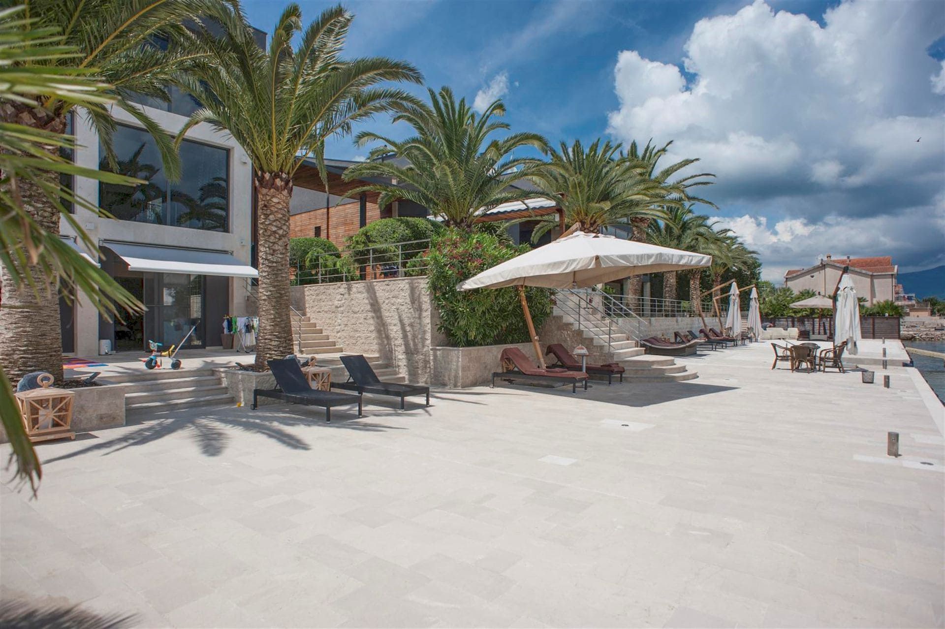 Terrace of a modern villa in Krašići, Montenegro: sun loungers, palm trees, and direct access to the sea from a private beach.