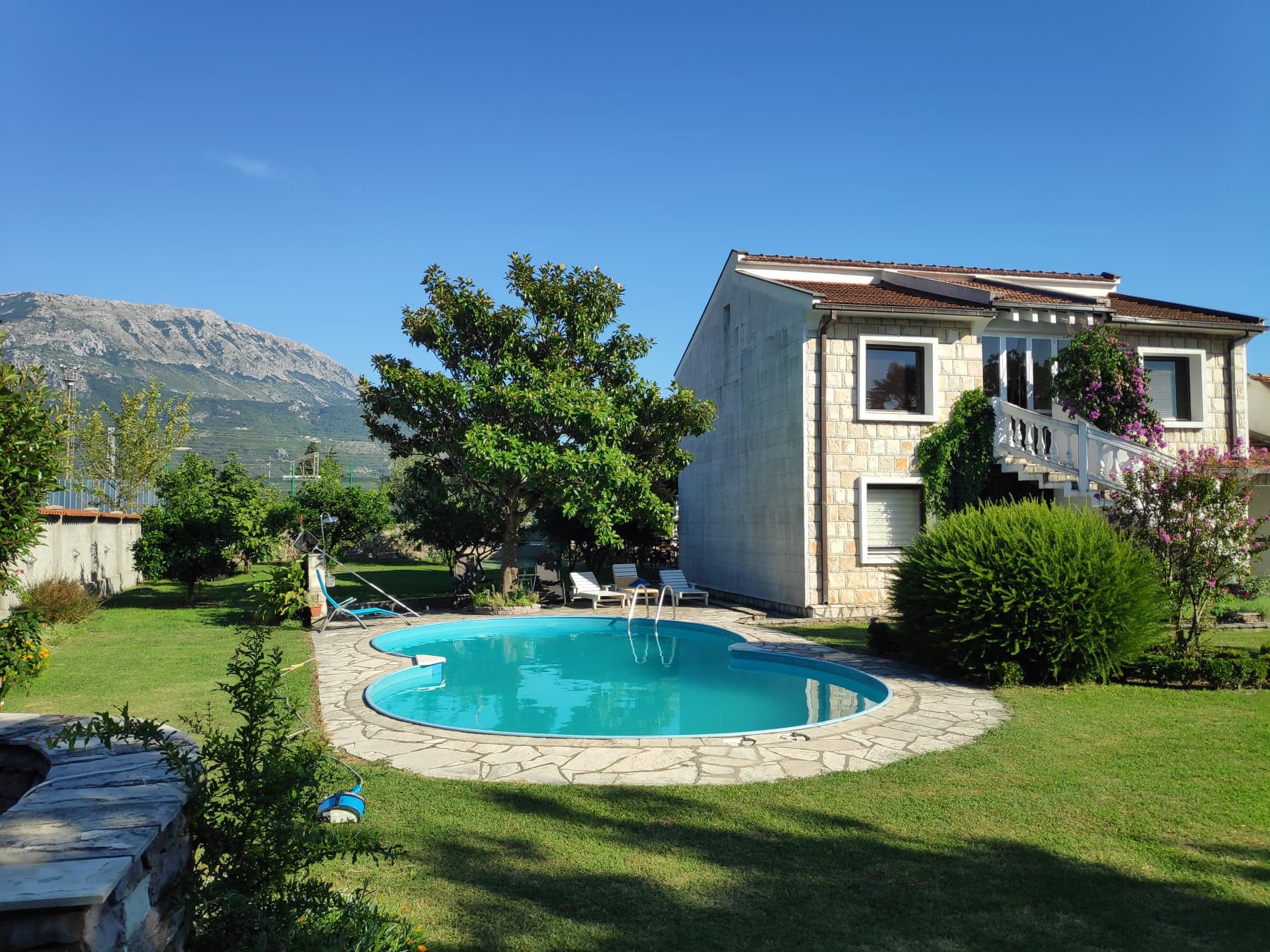 Two-story house with a pool and garden in Bar, Montenegro, against a mountain backdrop on a sunny day.