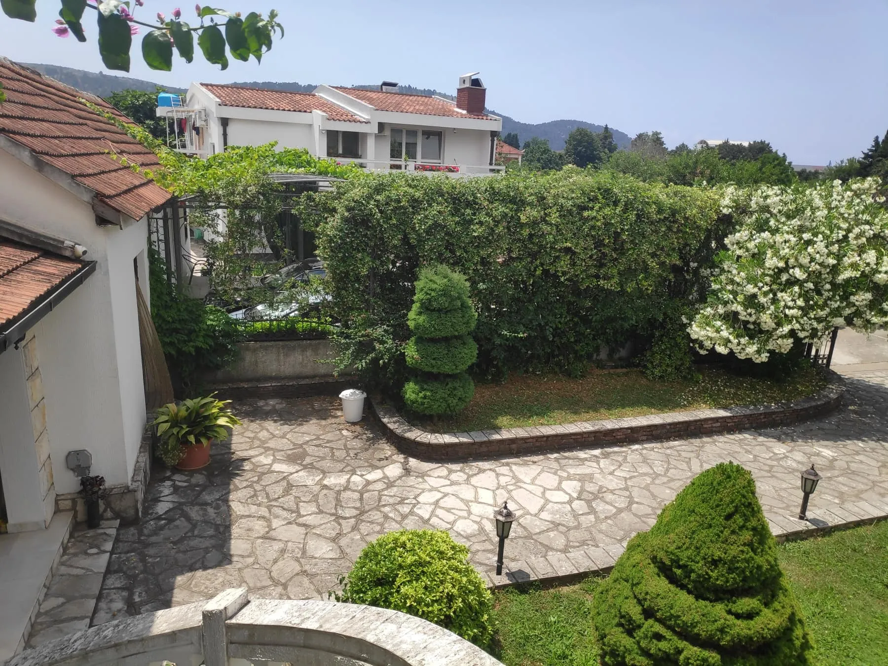 Stone courtyard with manicured bushes, trees, and a white house with a red tiled roof against mountains.