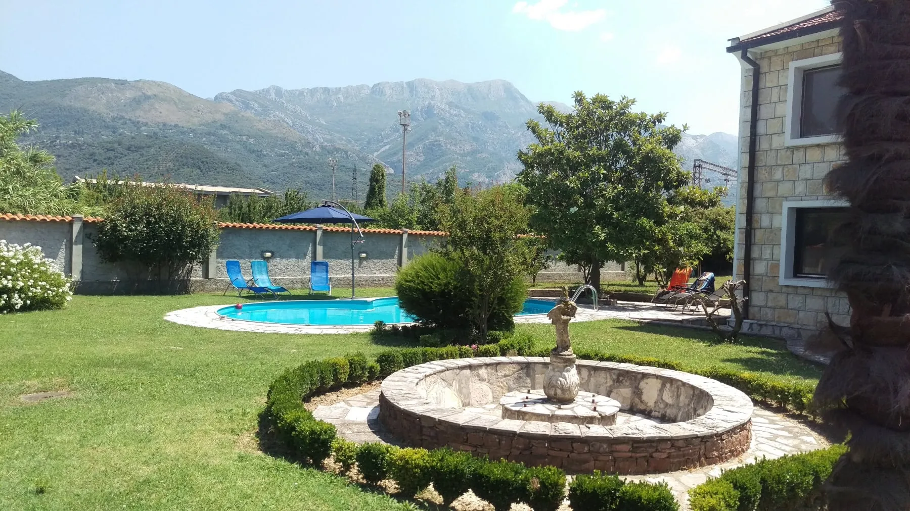 Garden with a swimming pool, stone fountain, lounge chairs, and mountains in the background.