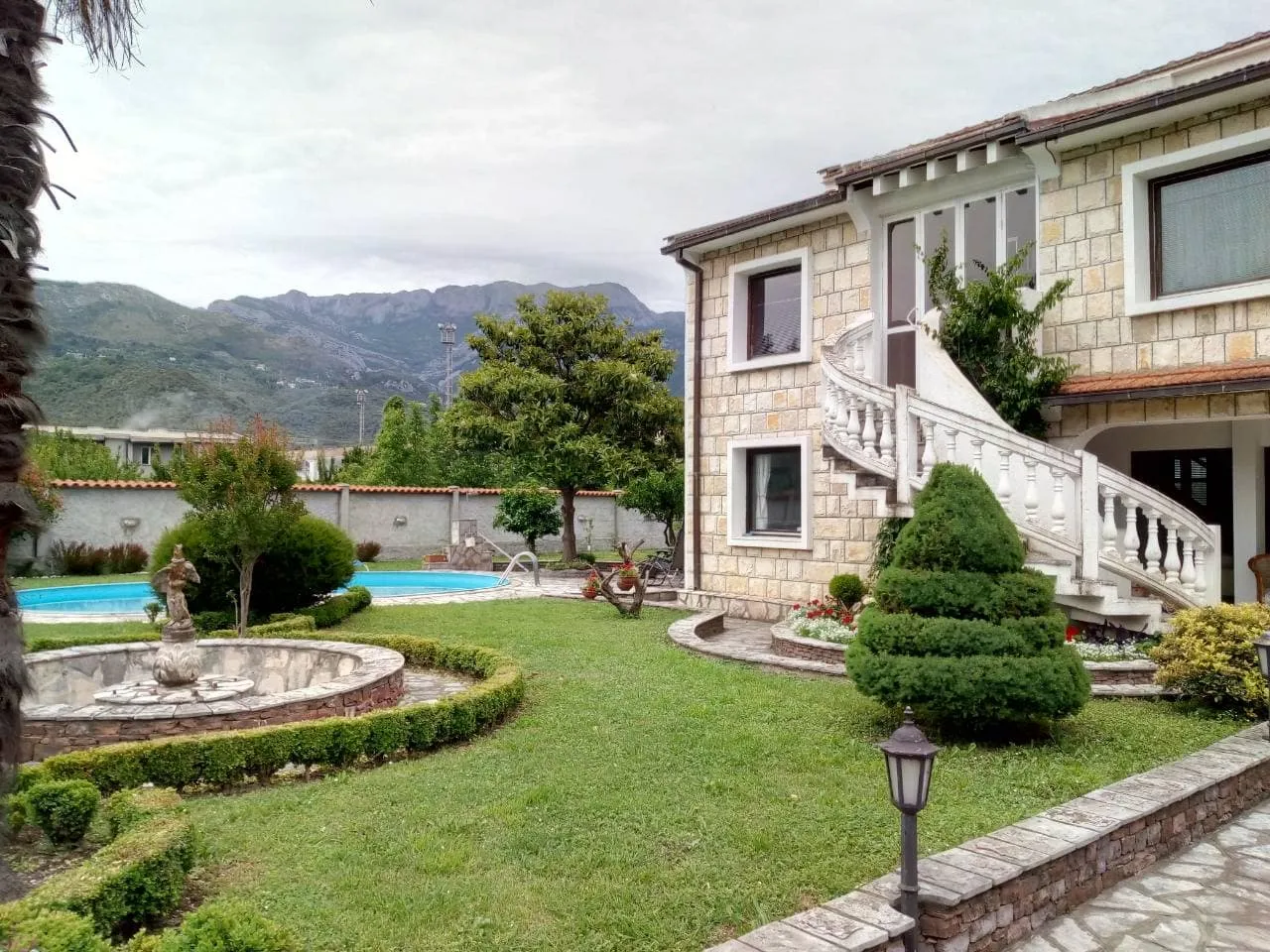 Stone house with white staircase, green lawn, fountain, and pool set against a mountain backdrop.