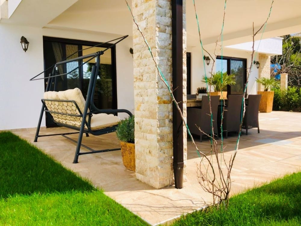 House terrace in Dobri Vody, Montenegro: garden swing, dining area, stone-clad columns, and a view of the garden.
