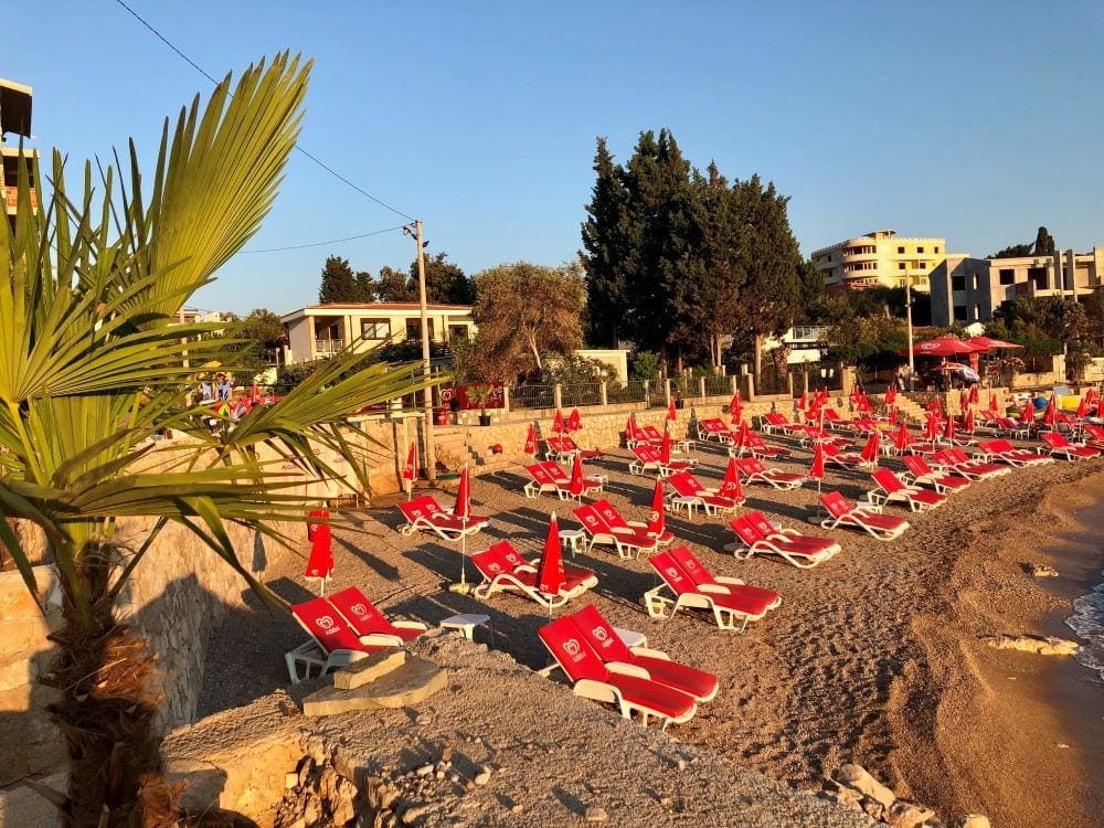 Beach with red sun loungers and a palm tree by the house in Dobri Vody, Montenegro: view of the seafront and infrastructure.