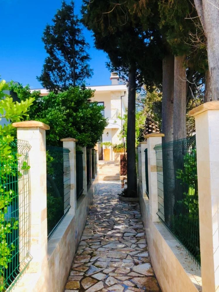 Stone path to the house in Dobri Vody, Montenegro, surrounded by green trees and a modern fence.