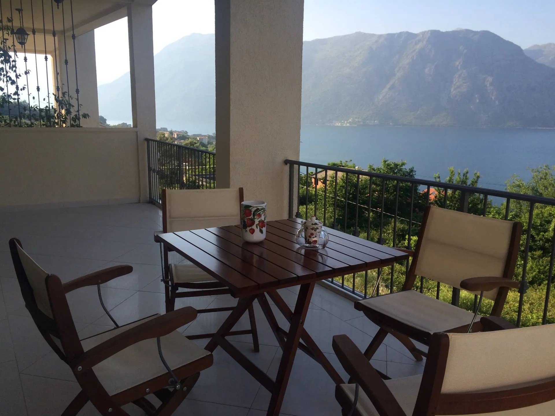 Balcony with wooden table and chairs overlooking the sea and mountains behind a metal railing.