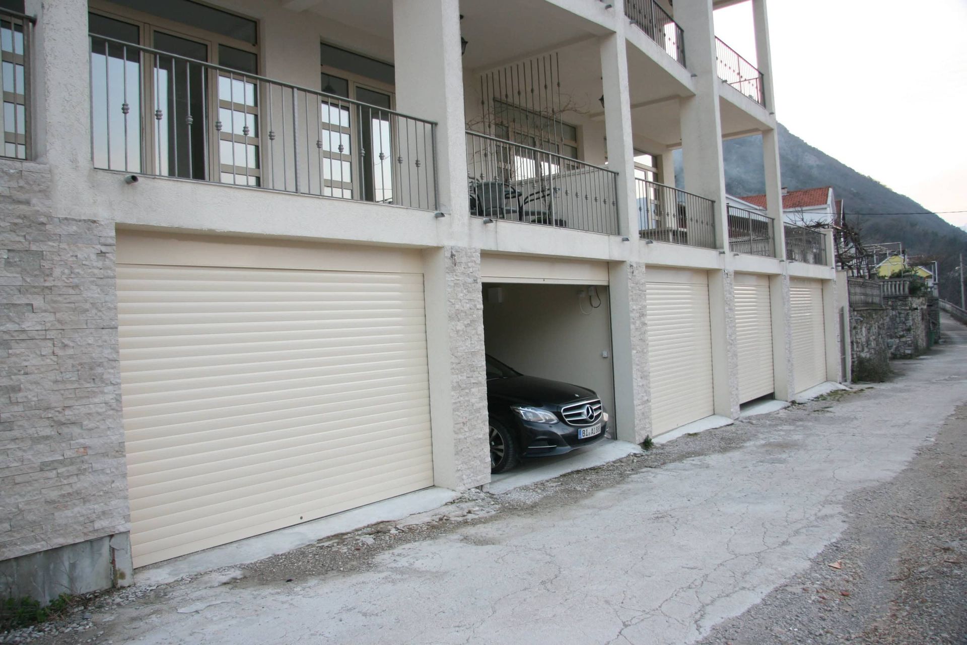 House facade in Prčanj, Montenegro: garage bays with roller shutters, parking space, and balconies with wrought-iron railings.