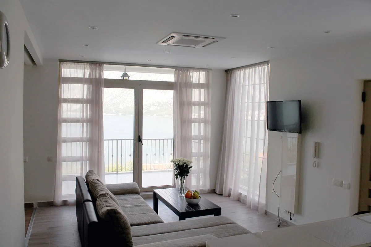 Bright room with a grey sofa, coffee table, and wall-mounted TV. Large glass doors lead to a balcony.