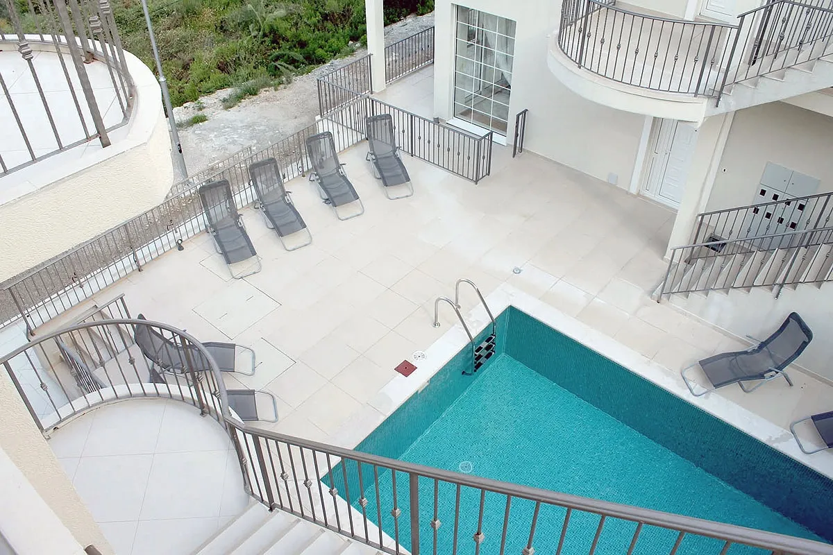 High-angle view of a turquoise swimming pool surrounded by a tiled terrace with lounge chairs and metal railings.