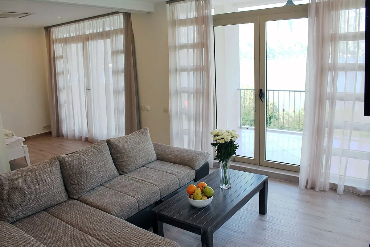 Living room with gray sofa, wooden table holding fruit. Balcony door and sheer curtains are visible.