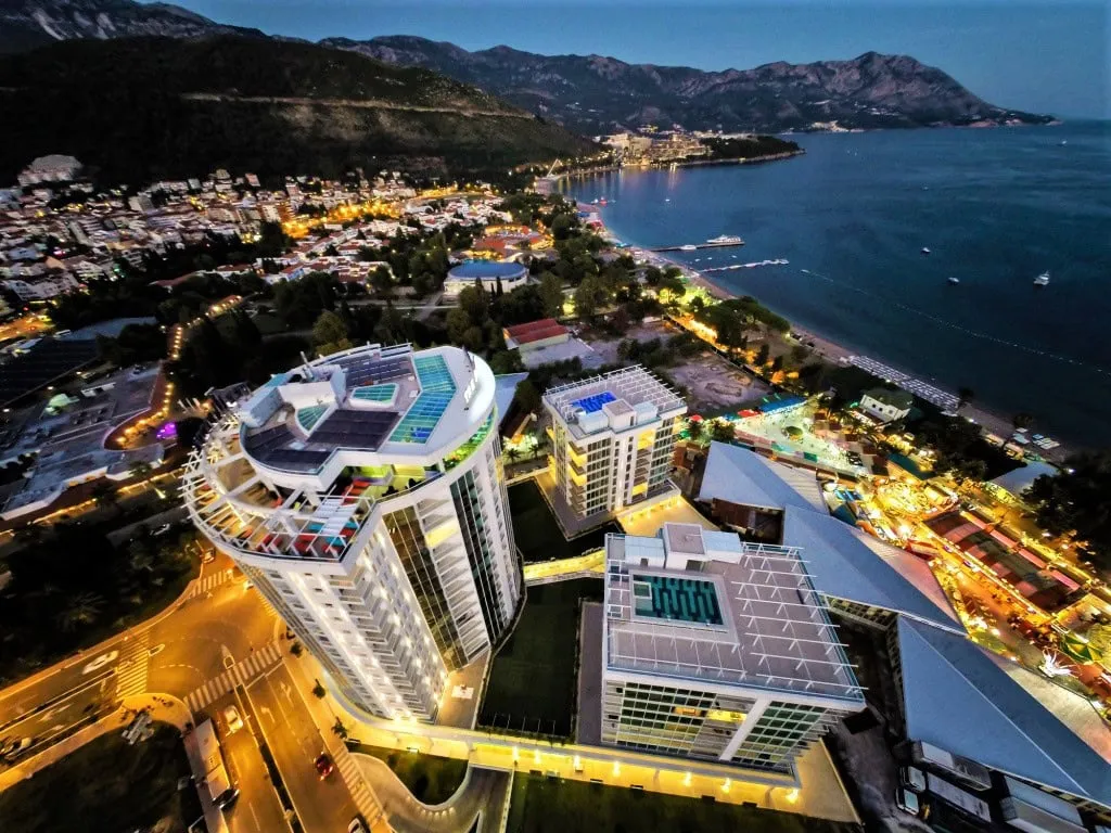 Aerial view of a coastal city with modern buildings, sea, and mountains at dusk.