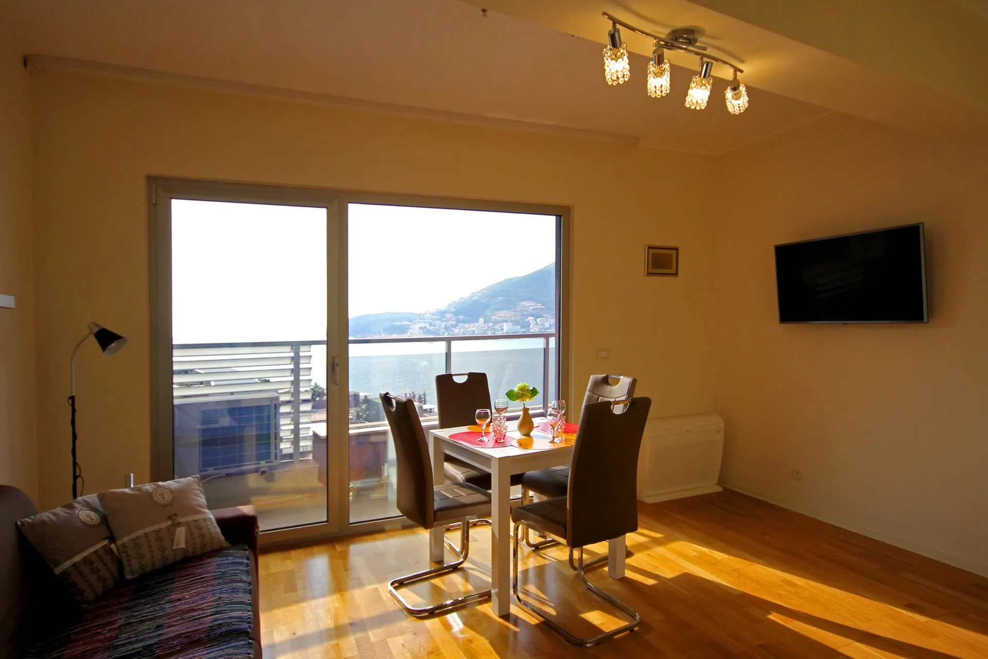 Living room with dining table, sofa, and wall-mounted TV. Features a large sliding glass door leading to a balcony with a sea view.