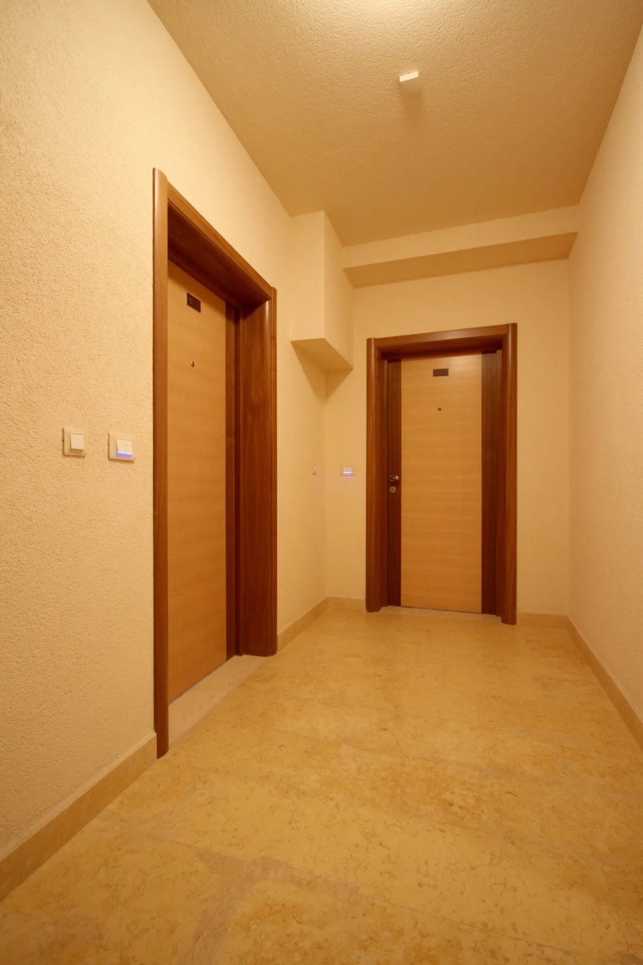 Indoor hallway with two wooden doors, textured walls, and beige tiled flooring.