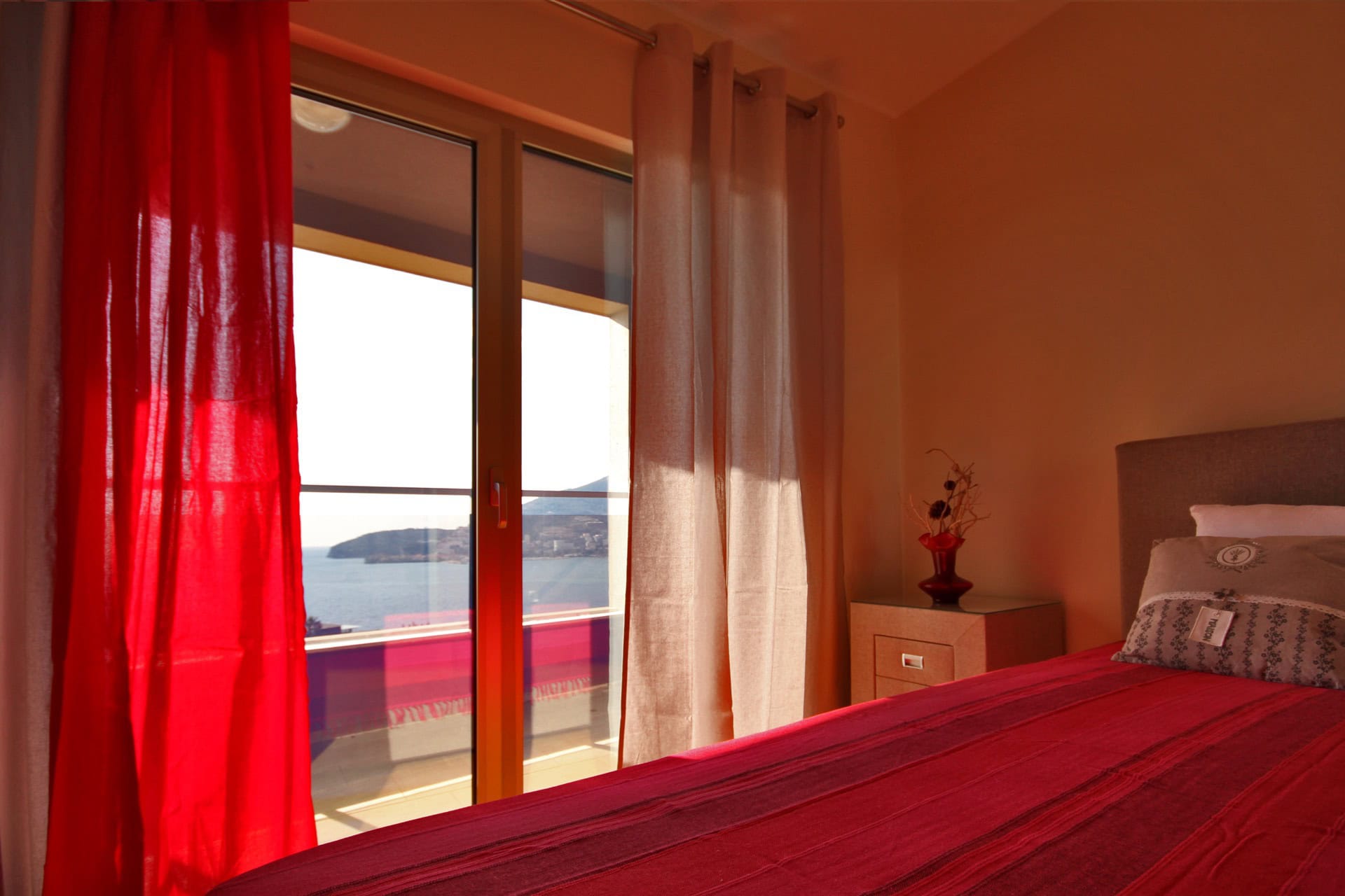 Bedroom in an apartment in Bečići, Montenegro: sea view from the balcony, red bedspread, bedside table, and panoramic window.