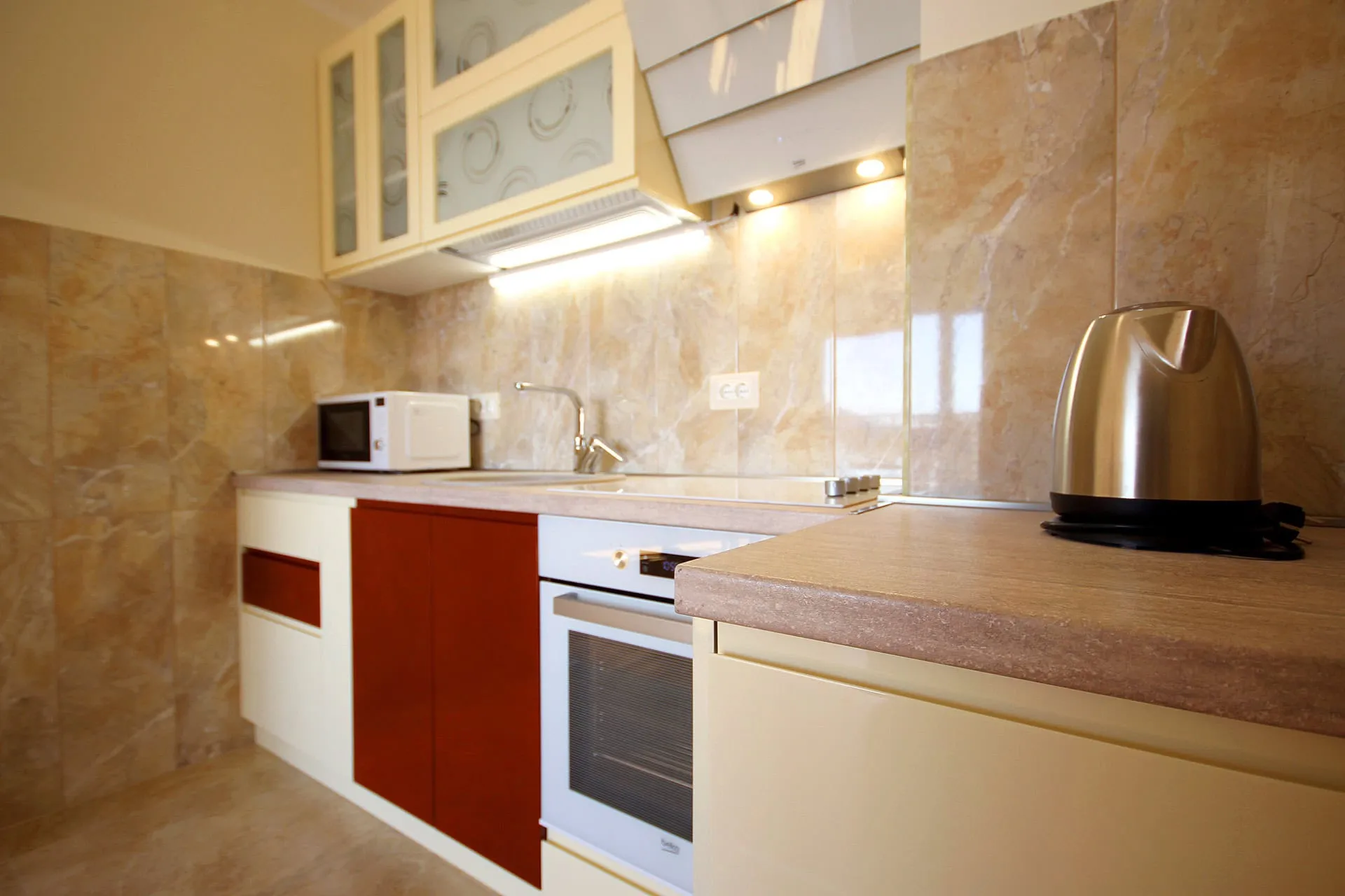 Kitchen with beige tiles, red and white cabinets, microwave, built-in oven, and a stainless steel electric kettle on the counter.