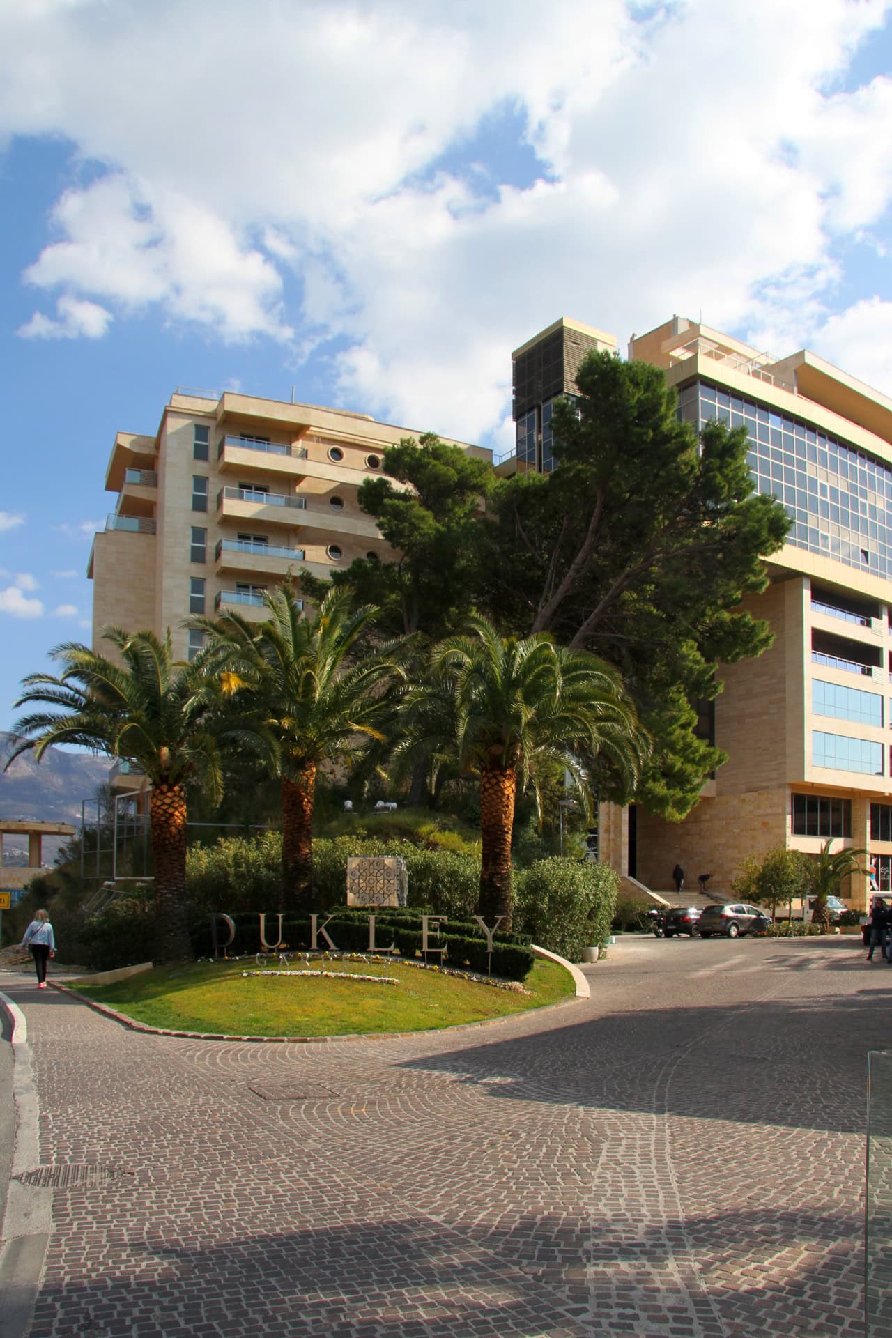Facade of the Dukley Gardens residential complex in Bečići, Montenegro: modern architecture, palm trees, and landscaped grounds.