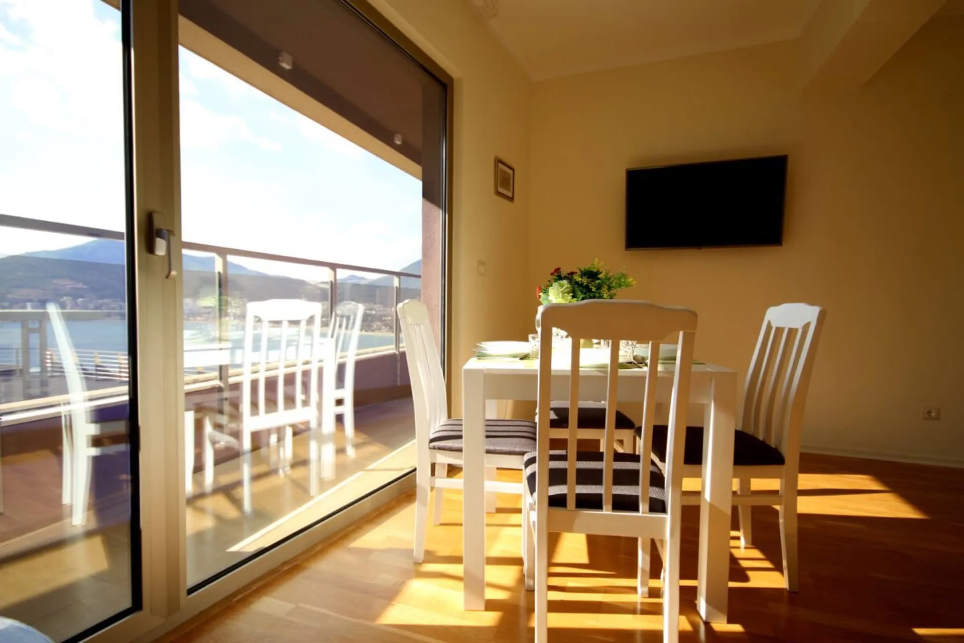 Bright room with a white dining table, chairs, and wall-mounted TV. Large glass door leads to a balcony overlooking water and mountains.