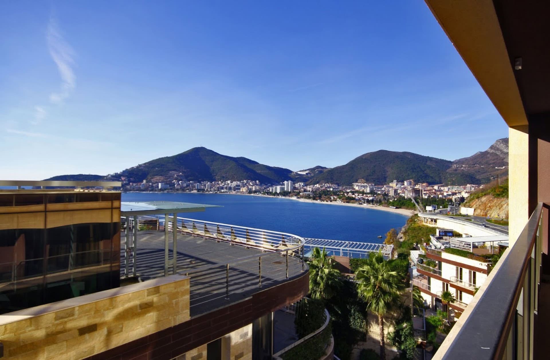 View from the apartment balcony in Bečići, Montenegro: a panorama of the Adriatic Sea, the beach, and the mountains on a sunny day.