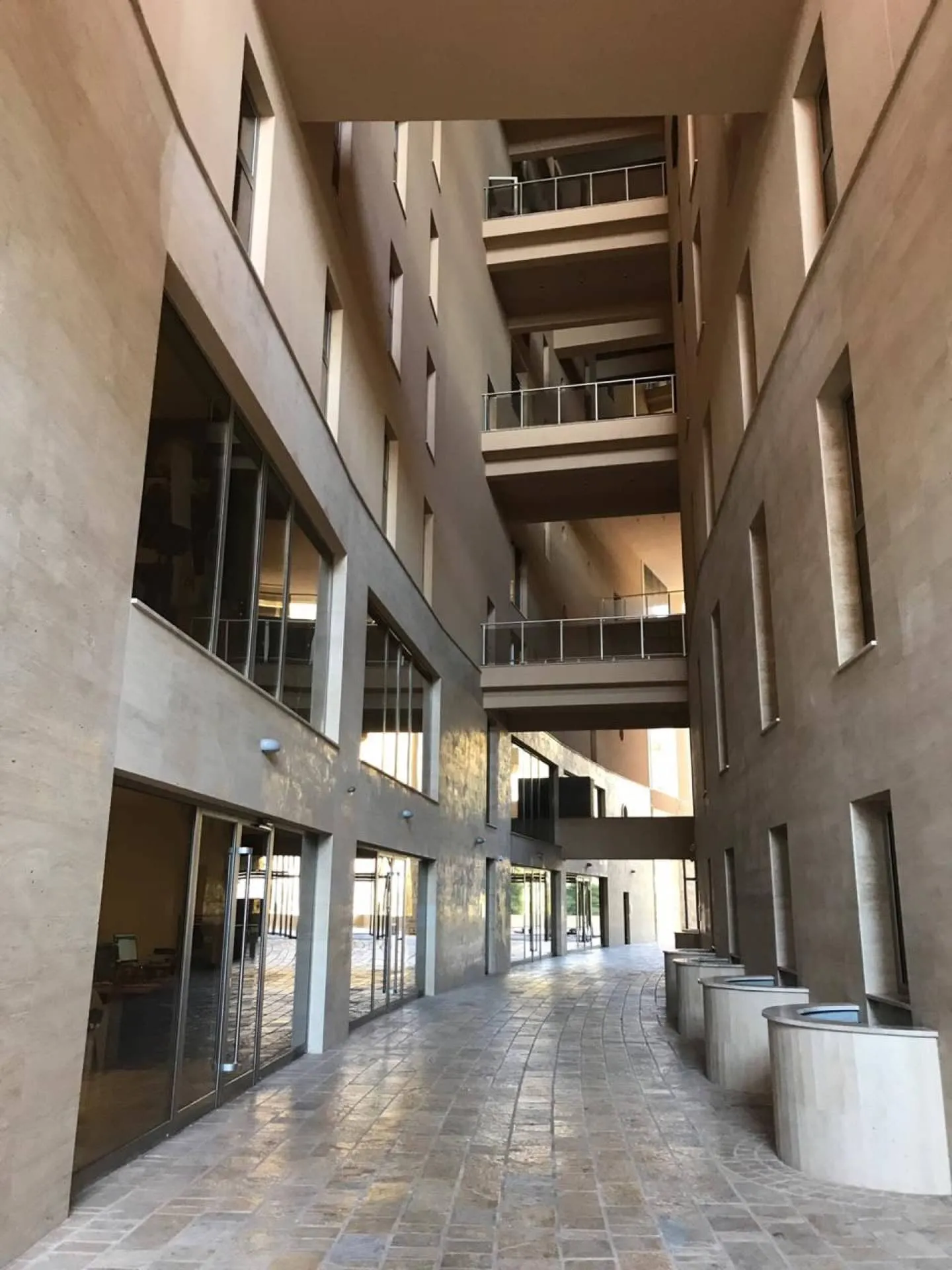 Long stone corridor with arched openings, upper balconies, and glass doors on the left side of the walkway.