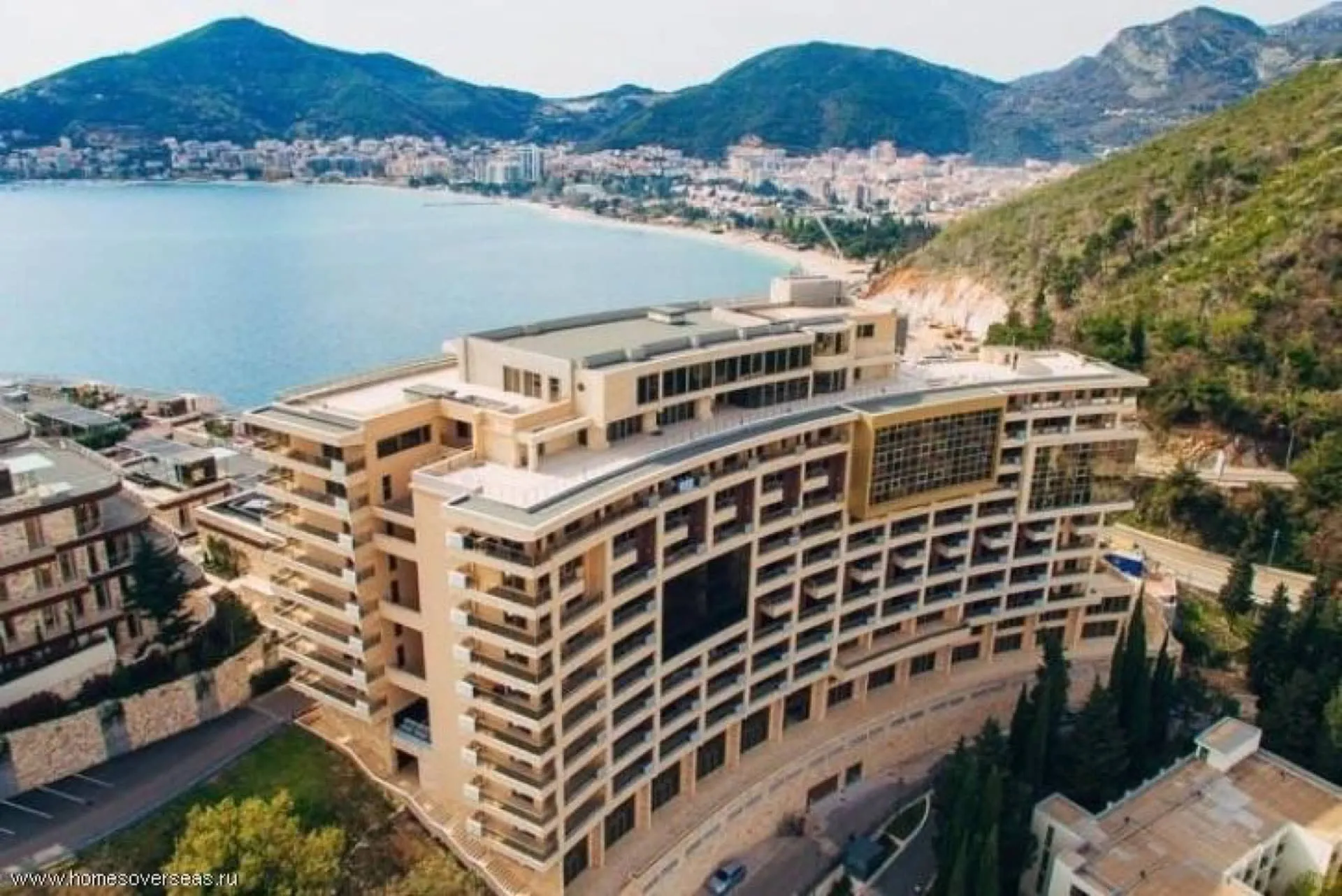 Aerial view of a large multi-story coastal building with mountains in the background and a cityscape along the bay.