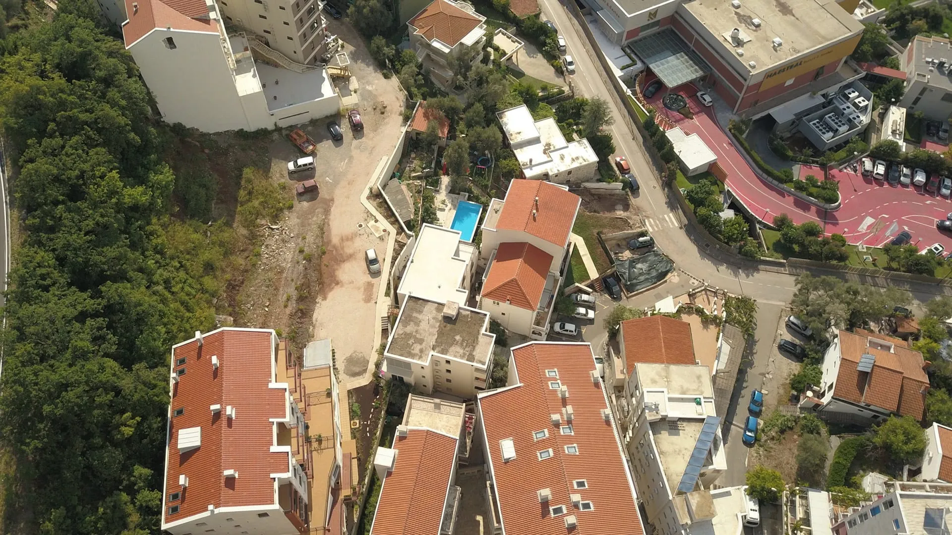 Aerial view of a residential area showing red-tiled rooftops, a road, trees, parked cars, and a small blue swimming pool.