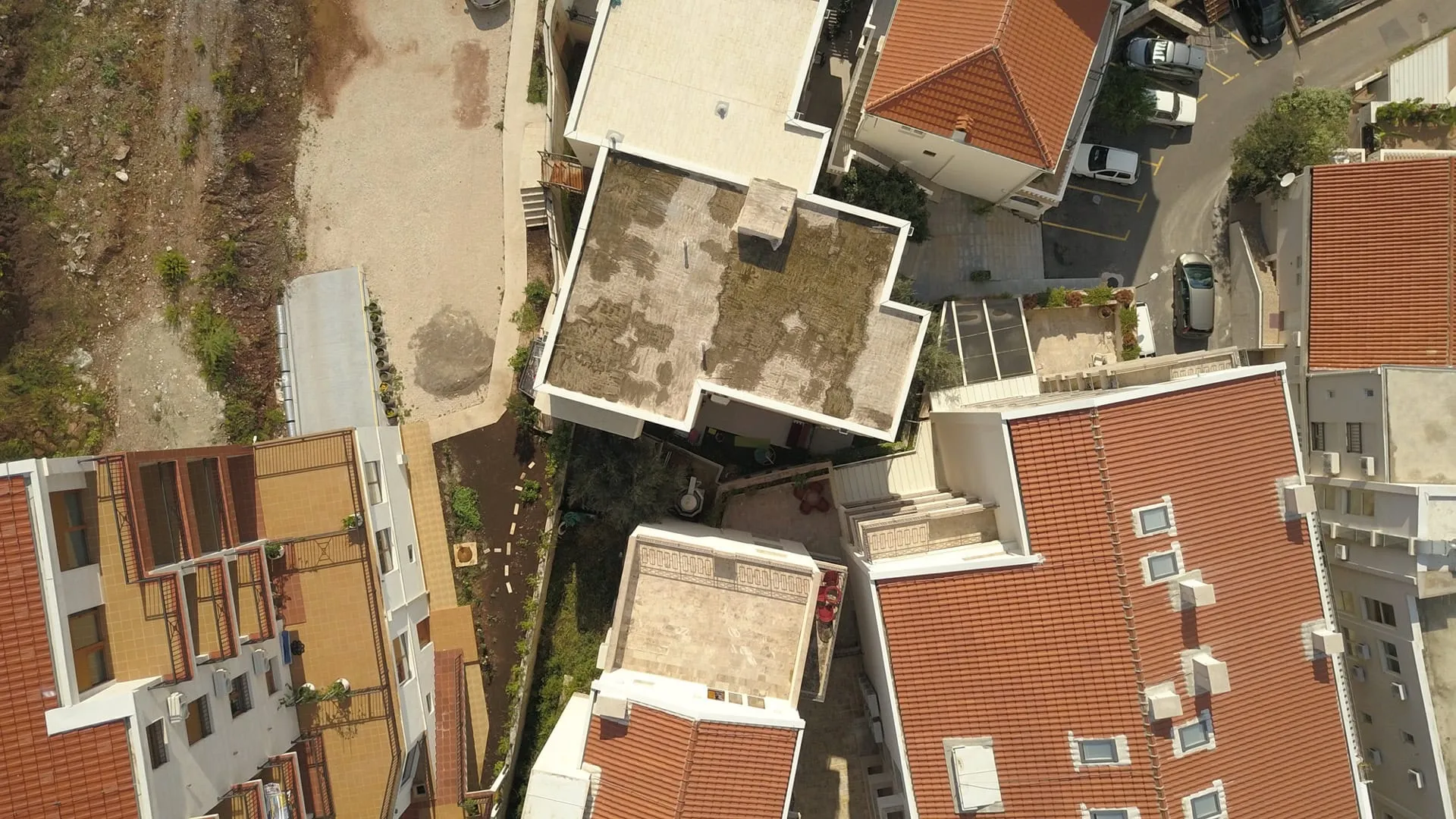 Aerial view of residential rooftops with red tiles, flat terraces, and adjacent parking areas with cars.
