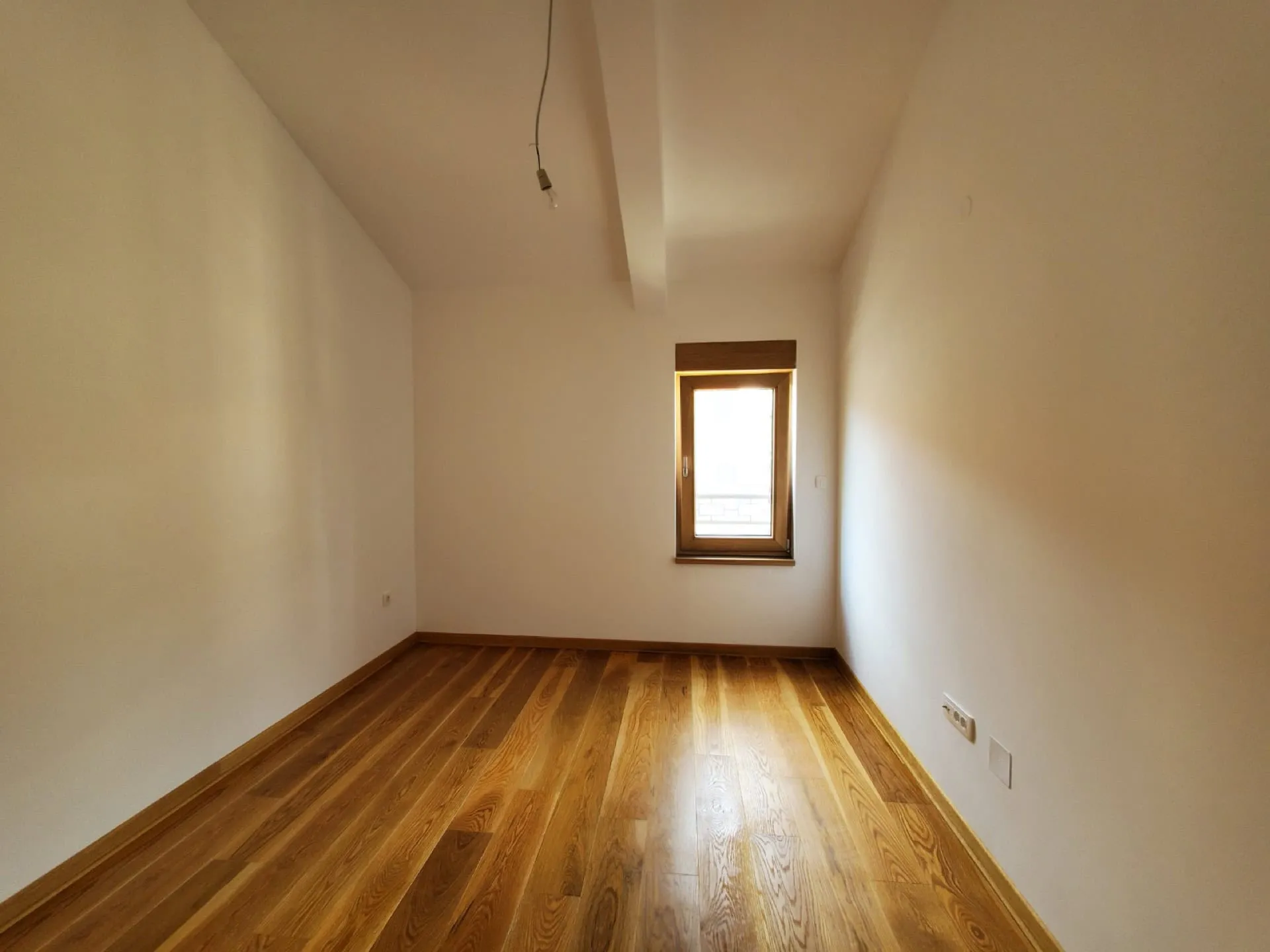 Empty room with wooden flooring, white walls, a small window, and a single bare bulb hanging from the ceiling.