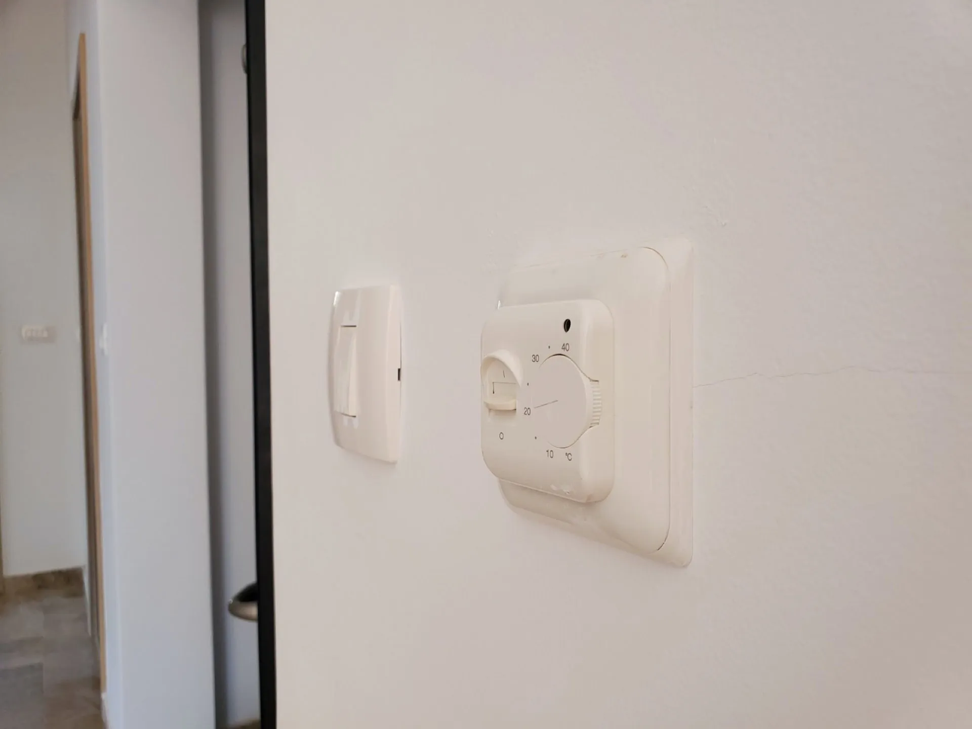 White thermostat and light switch on a wall with a visible crack; a doorway to a hallway is seen on the left.