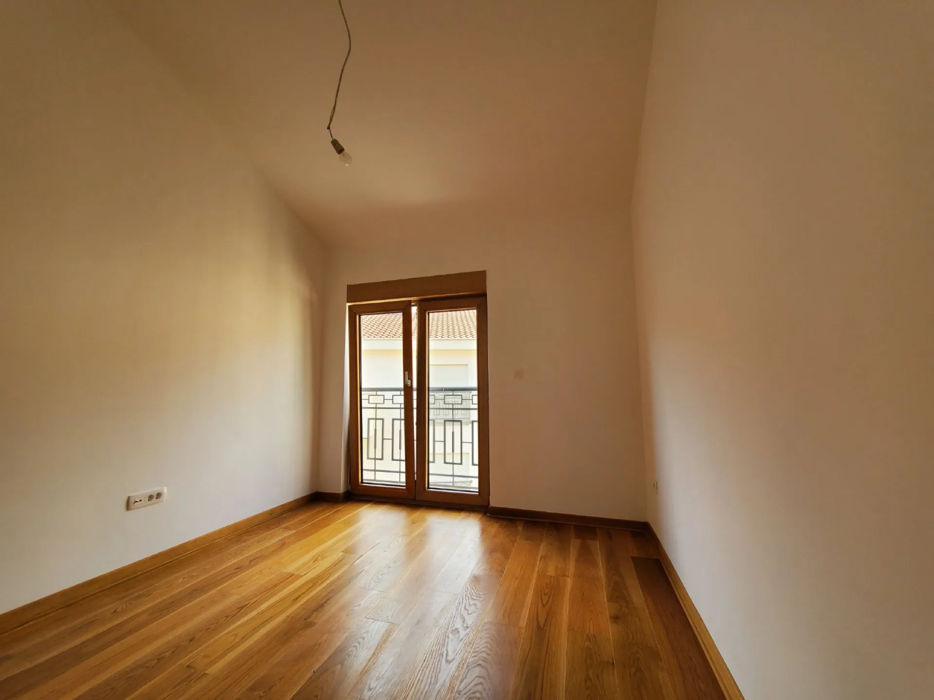 Empty room with wooden flooring and balcony door, hanging bulb, white walls, no furniture.