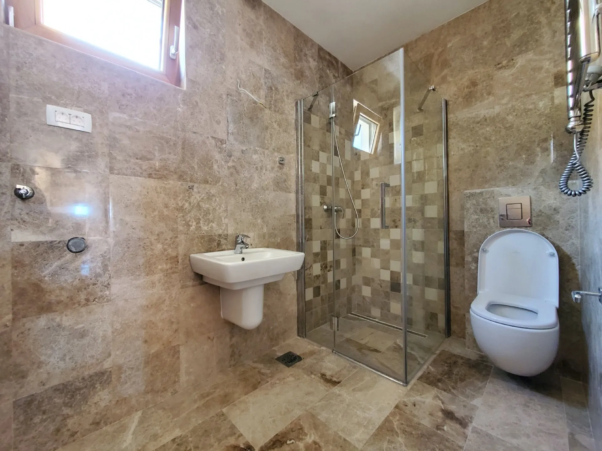 Bathroom with marble tiles, glass shower enclosure, wall-mounted toilet, and pedestal sink.
