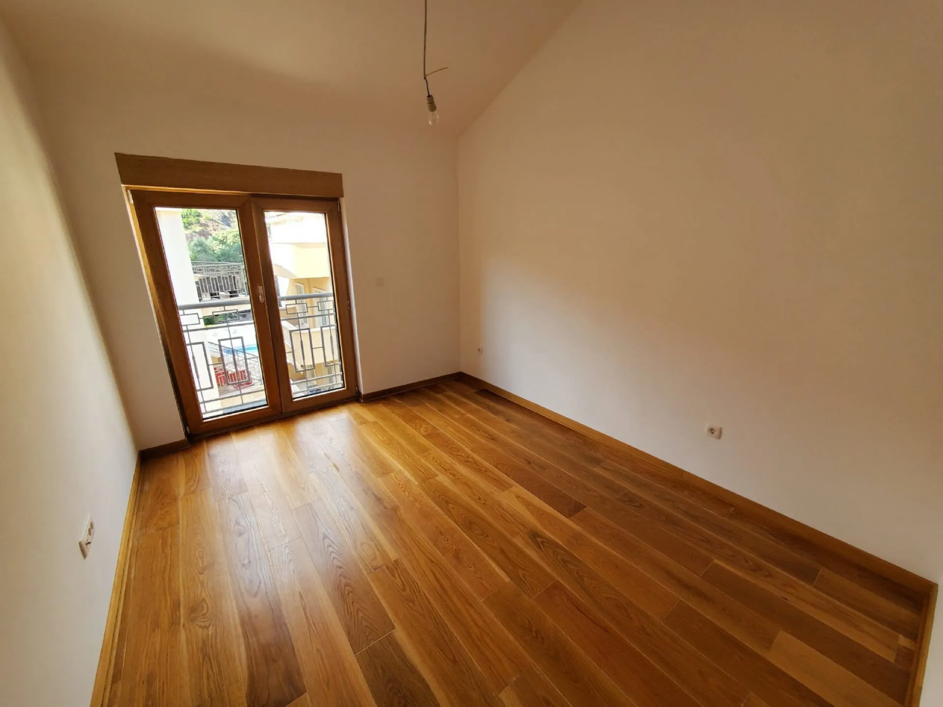 Empty room with wooden flooring and balcony access. No furniture, visible hanging light bulb.