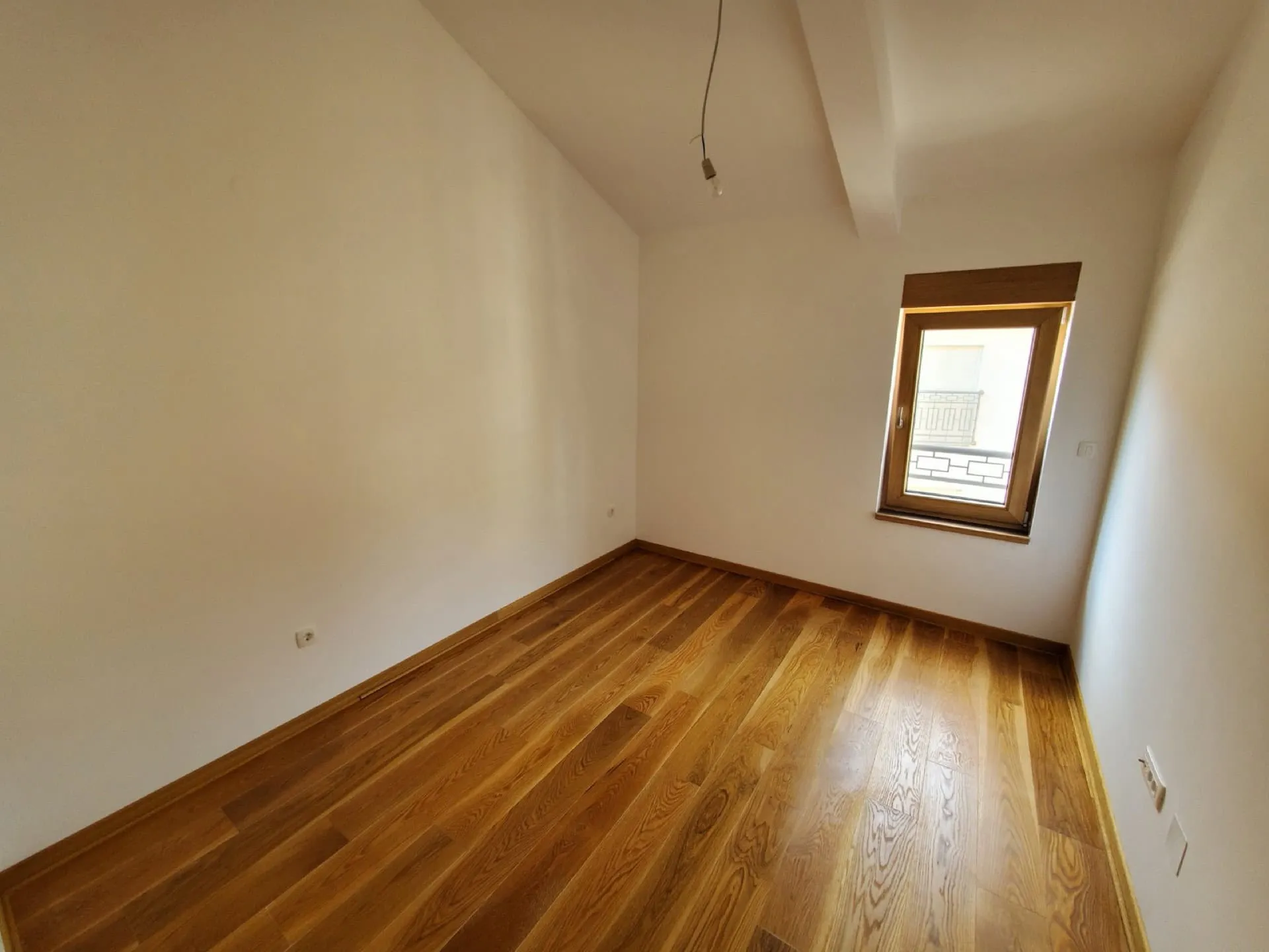 Empty room with wooden flooring and white walls, featuring a hanging bulb and a window.