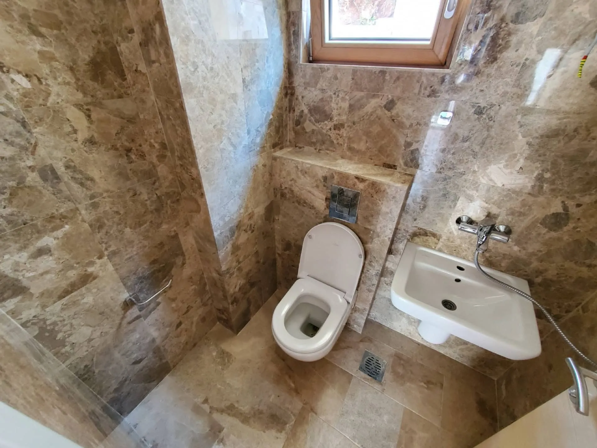 Bathroom with marble tiles, white toilet, bidet with shower hose, and a small window.
