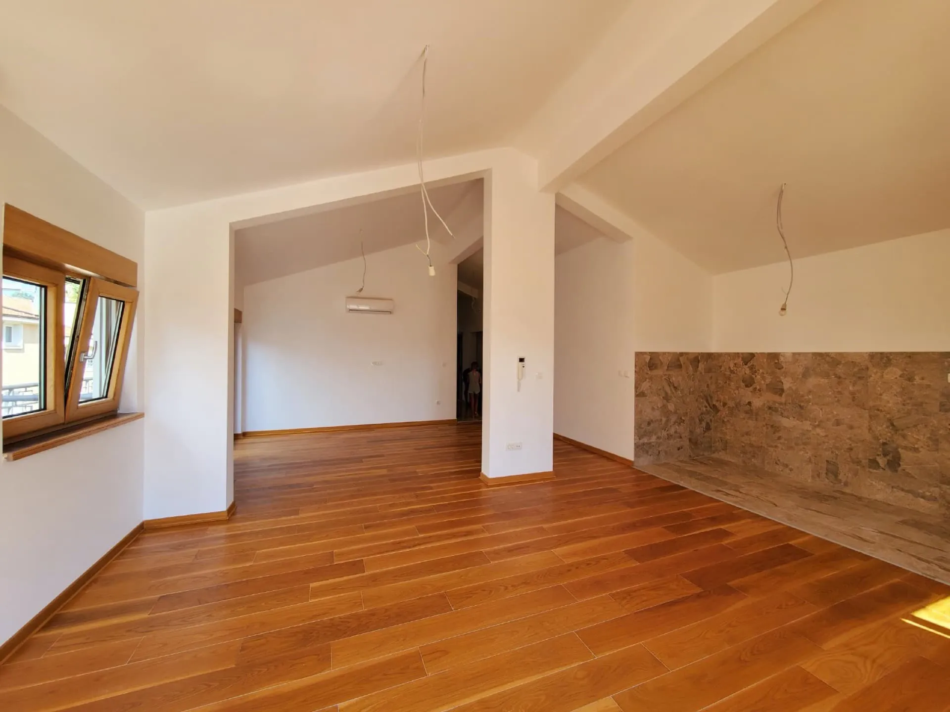 Empty room with wooden flooring, stone wall section, hanging wires, and a window looking outside.