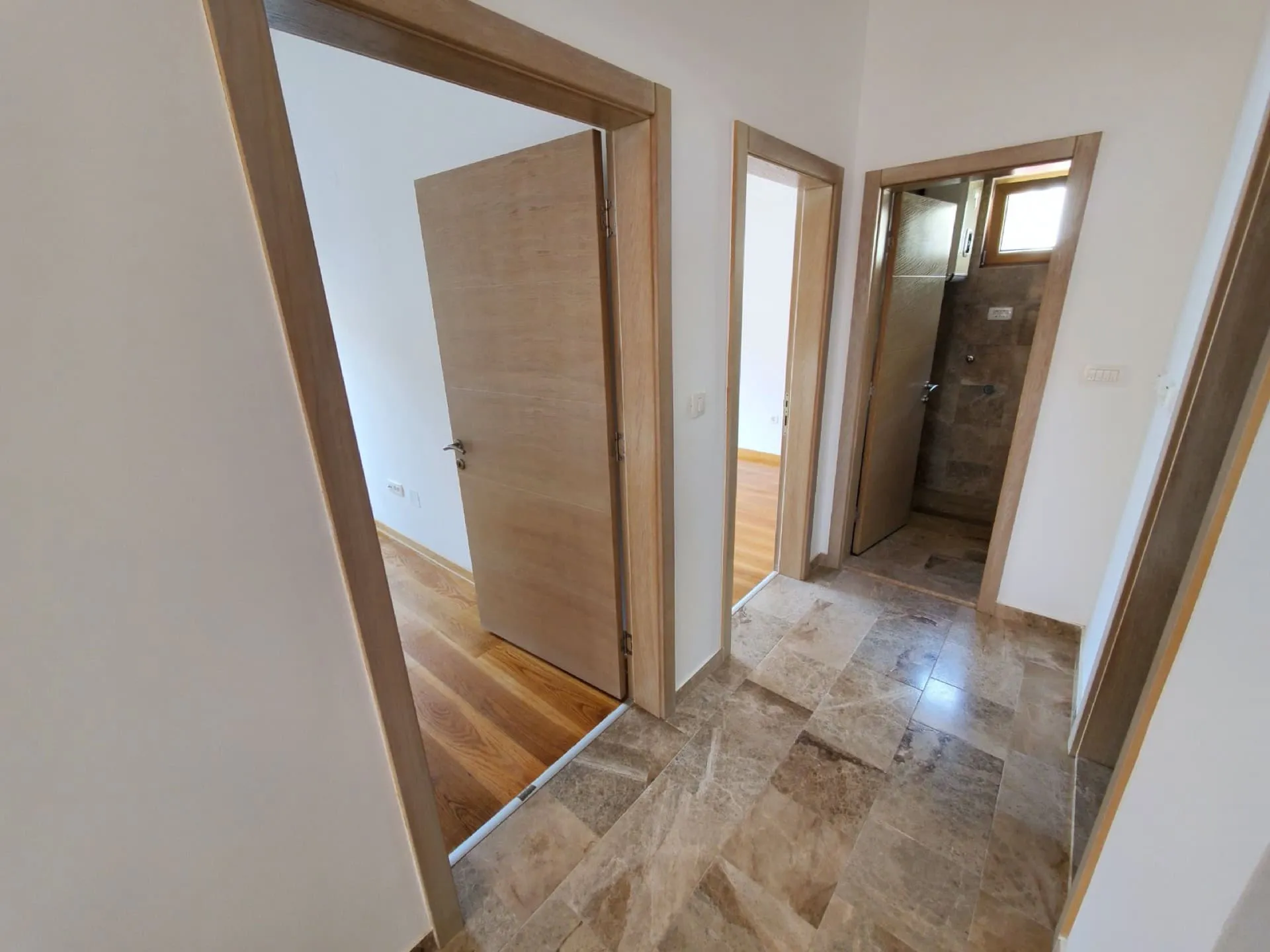 Hallway with stone tile flooring leading to rooms with wood floors and a bathroom with a glass door.