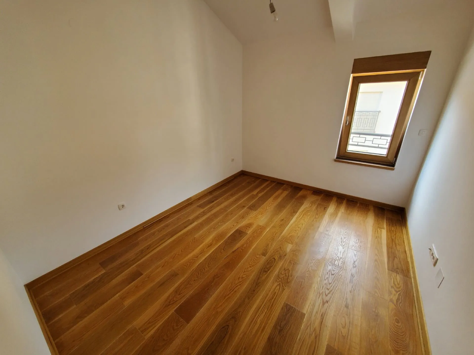 Empty room with wooden flooring, white walls, and a window overlooking a balcony railing.