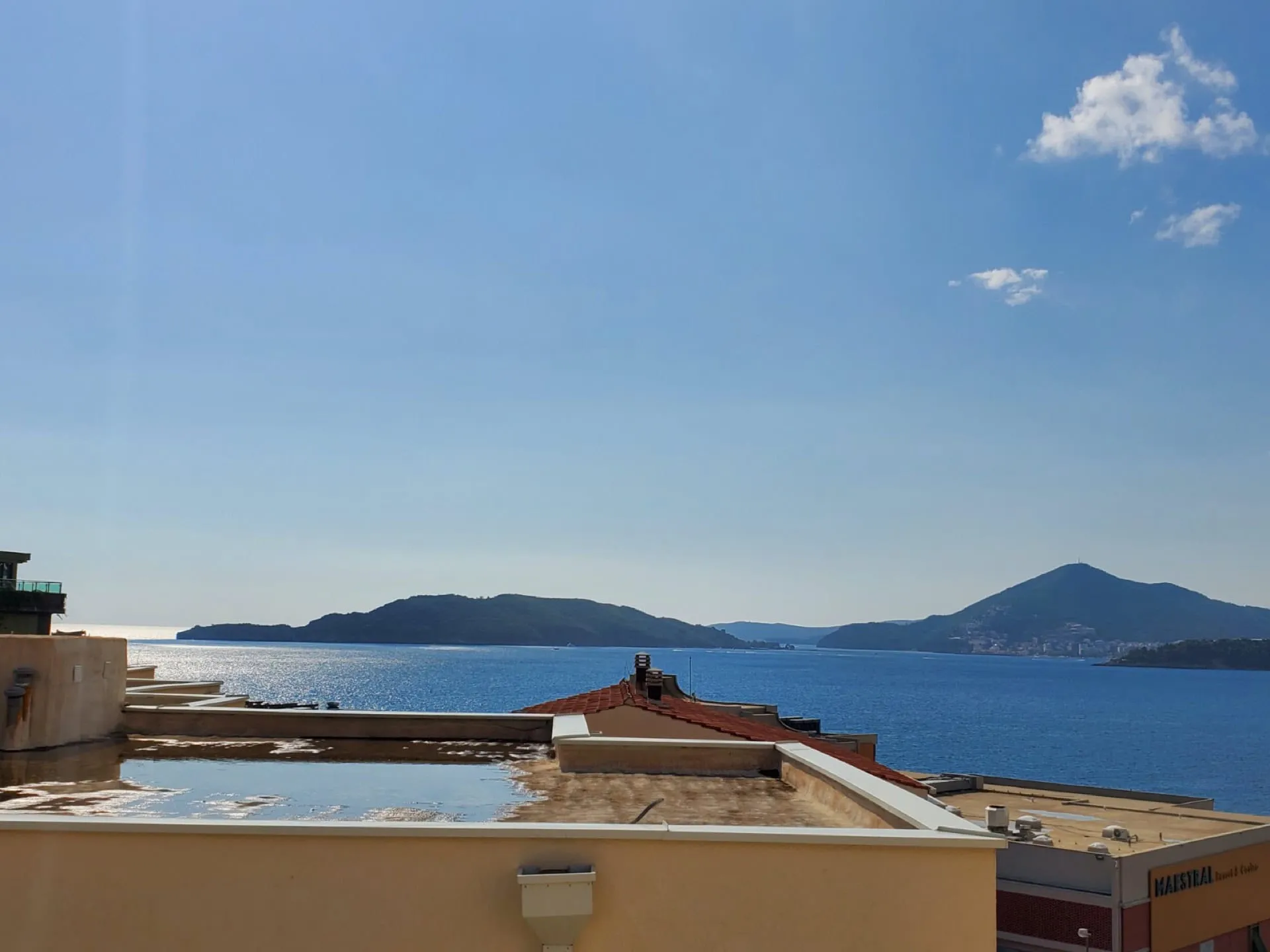 Rooftop view of the sea and islands under a blue sky, with parts of adjacent buildings visible.