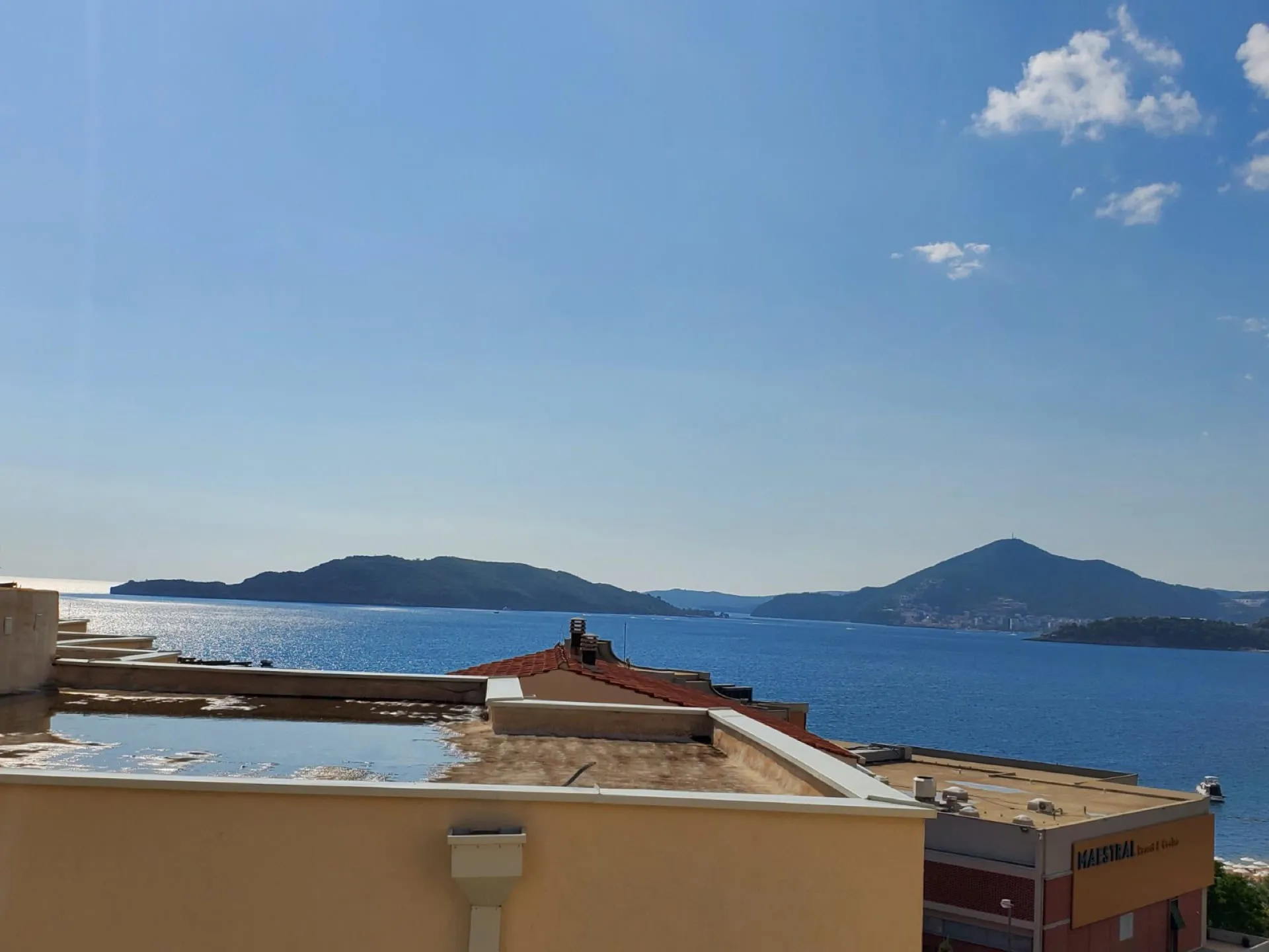 View from a rooftop overlooking the sea and islands under a blue sky. Parts of neighboring buildings and tiled roofs are visible.