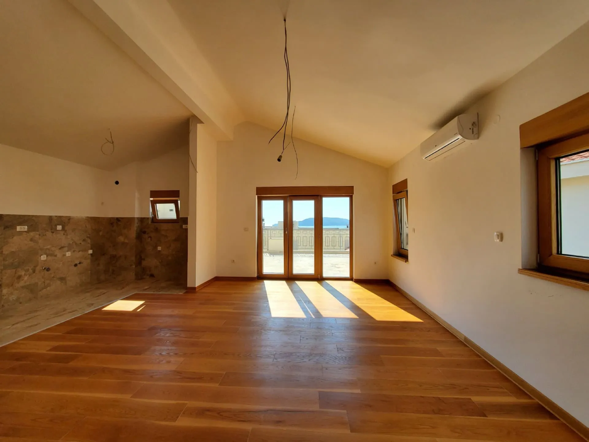 Empty room with wooden flooring, high ceiling, and balcony access. Features AC unit and unfinished tiled wall section.