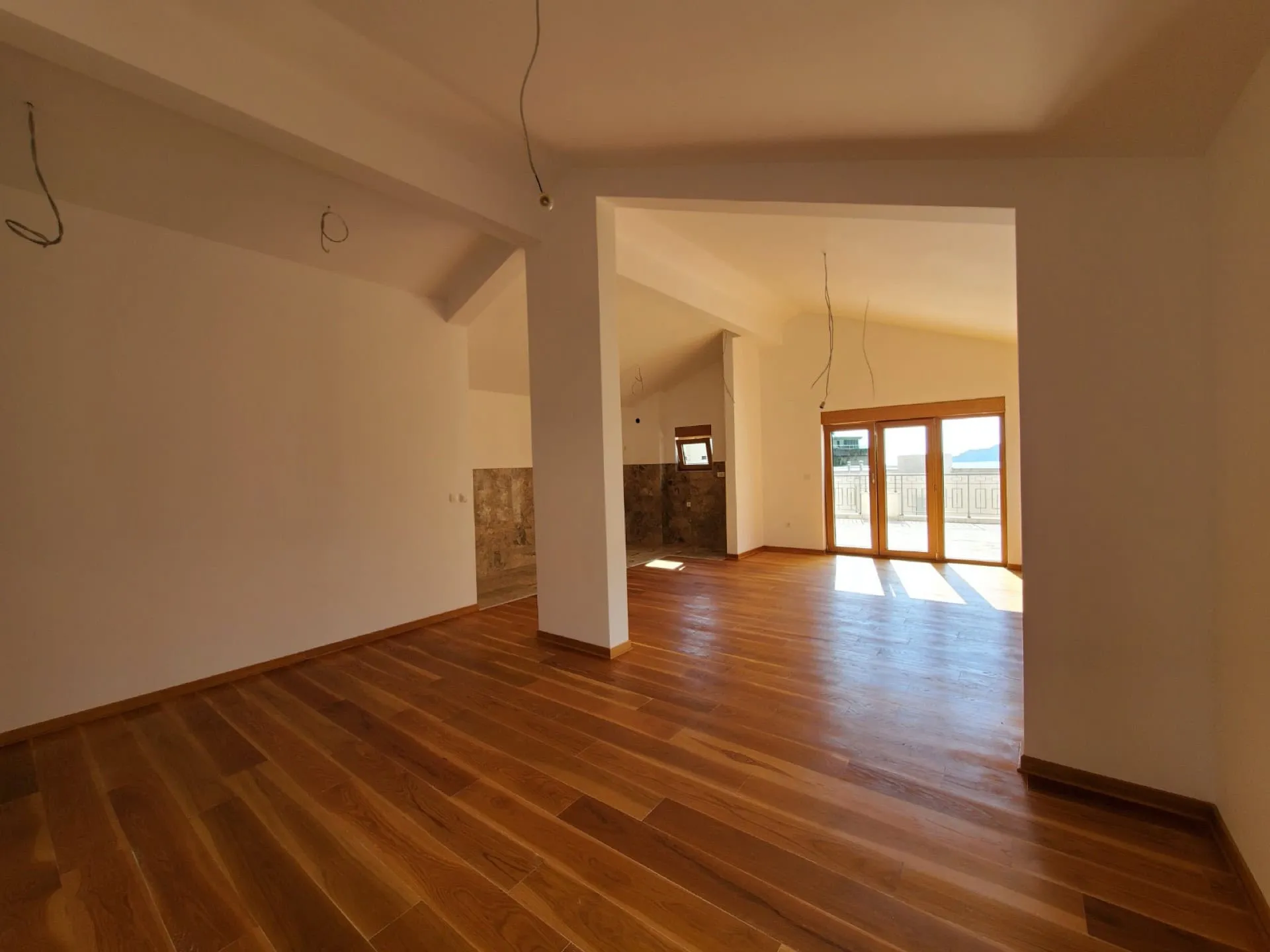 Empty room with wooden flooring, white walls, hanging wires, and a doorway leading to a balcony.