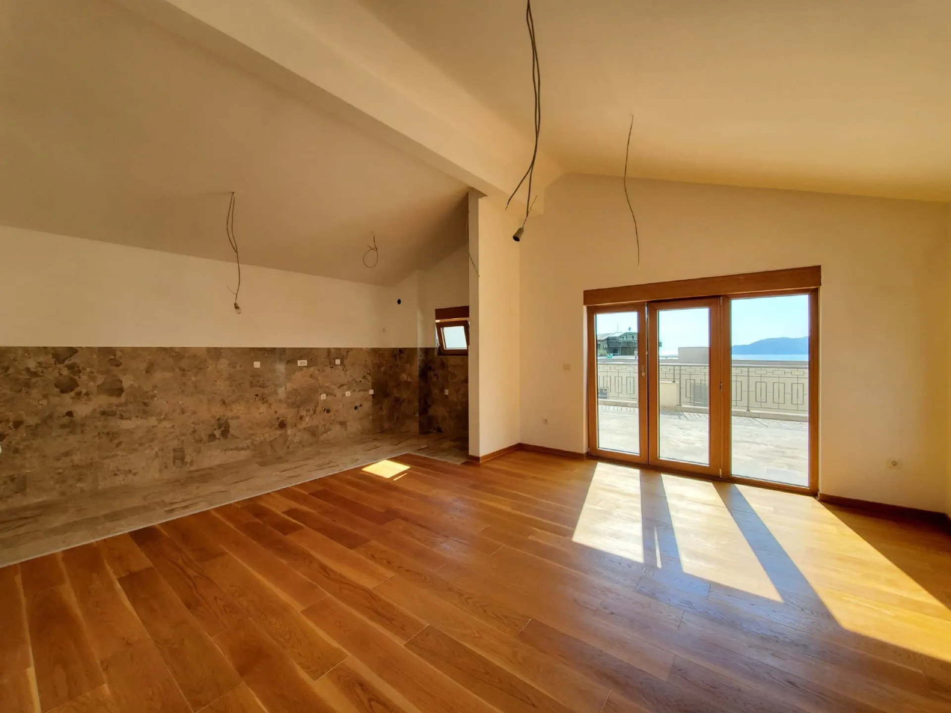 Empty room with wooden flooring and stone wall section, glass doors leading to a balcony.